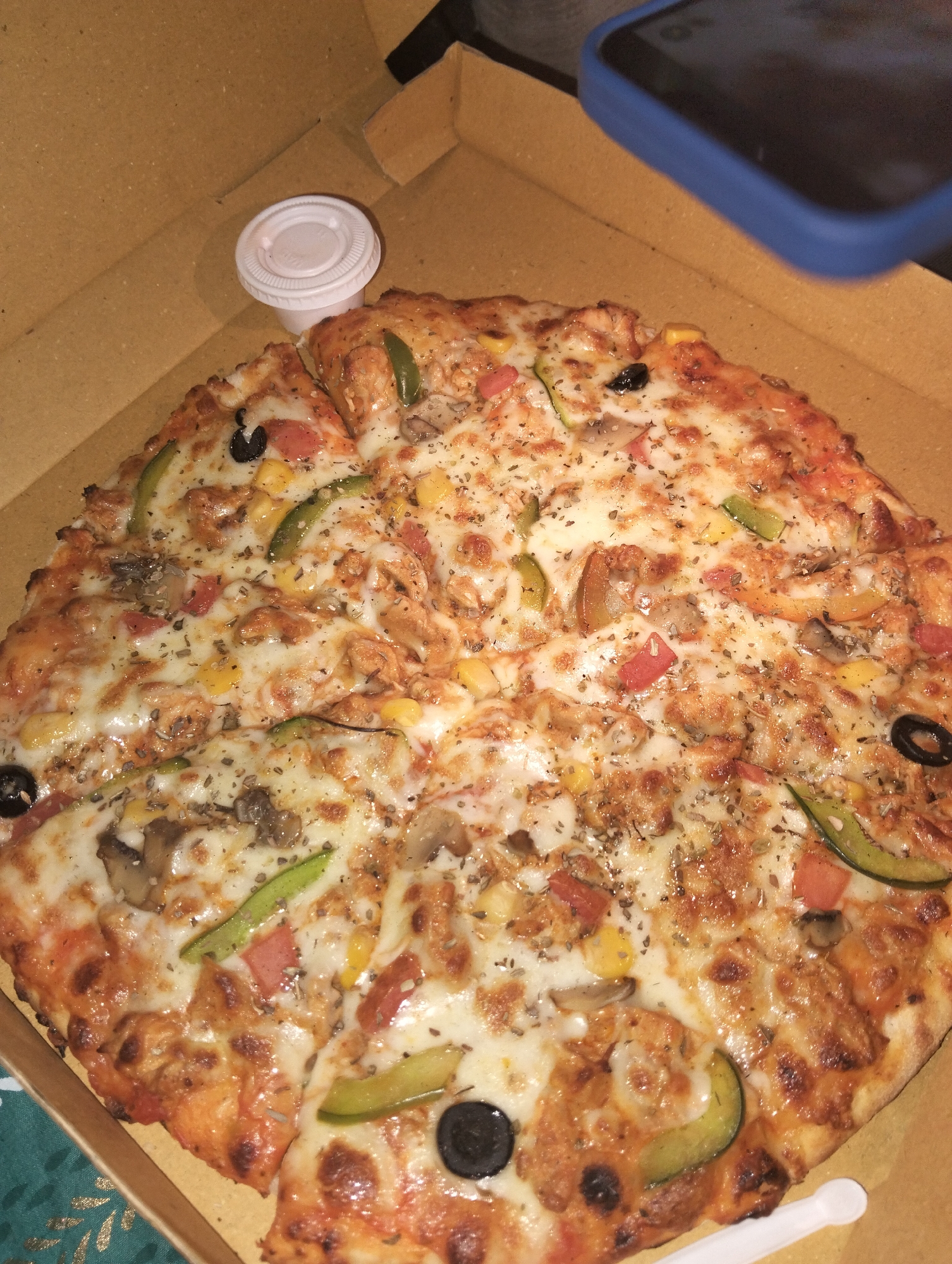 A top-down shot shows a pizza in a cardboard box, presumably for delivery or takeout. The pizza is round, cut into slices, and topped with melted cheese, what appear to be mushrooms, bell peppers, tomatoes, corn, olives, and some type of shredded meat or protein. A small, white, plastic container with a lid rests near the pizza slices, possibly containing sauce. A blue phone case is partially visible in the upper right corner of the image. The location is Dhaka, Bangladesh but there are no elements in the image to confirm or deny this location. No people are directly visible.