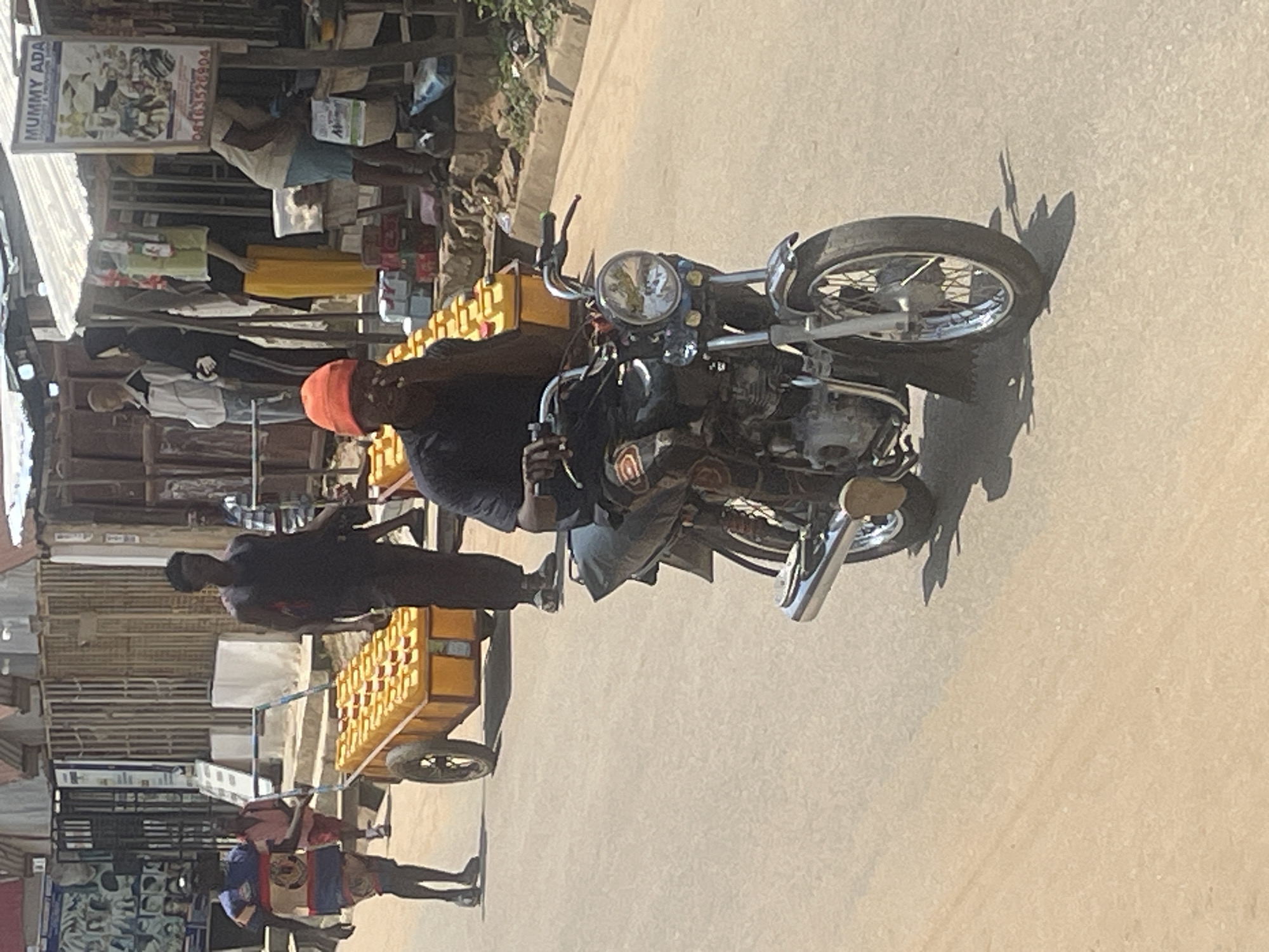 In Abuja, Nigeria, on a sun-drenched, paved street, a man in a black t-shirt and an orange beanie rides a motorcycle, his right hand on the handlebar and his left hand touching his face. The motorcycle, a dark color with chrome accents, has a visible front wheel with spokes and a silver exhaust pipe. Behind him, a cart loaded with yellow containers is being pushed by a man in a black shirt and dark pants. Another man in a blue and red striped shirt and patterned shorts is also present near the cart, his hands by his side. In the background, a market or shop area is visible with various goods and mannequins on display, and a sign with text, one of which reads "MUMMY ADA". The environment appears to be an outdoor urban setting with buildings and street vendors.