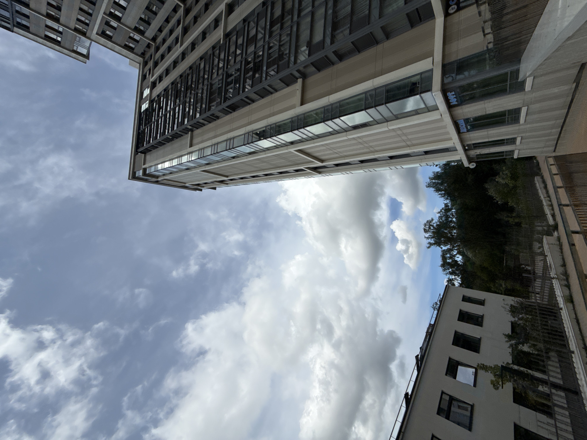 The image depicts a street-level view of a modern, multi-story residential building in Moscow, Russia, under a partly cloudy sky. The building's facade is characterized by a combination of beige and dark gray panels, with numerous large windows. The ground floor features storefronts with visible signage, including "ARTICHM" and "LOFT." To the left, a lower, white building with a sloped roof and multiple windows is visible, partially obscured by trees. The scene appears to be a residential area with some commercial spaces integrated into the ground floor of the taller building. The overall impression is of contemporary urban architecture.