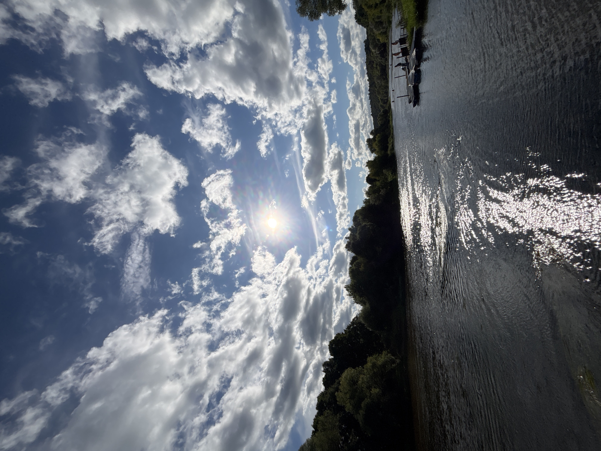 The image depicts a wide, dark river flowing through a verdant landscape. The sun, partially obscured by dramatic, puffy white clouds against a bright blue sky, casts a dazzling reflection on the water's surface, creating streaks of shimmering light. Along the left bank, a dense line of dark green trees forms a lush border, casting some of the foreground water into shadow. On the right bank, a wooden dock with several small boats moored to it is visible. The boats are white with dark accents and appear to be small recreational vessels. The overall impression is of a tranquil, sunlit day on a river, likely in a rural or semi-rural setting. The specific location, Zvenigorod, Russia, suggests a scene that could be typical of the Moscow Oblast region, known for its rivers and natural beauty.