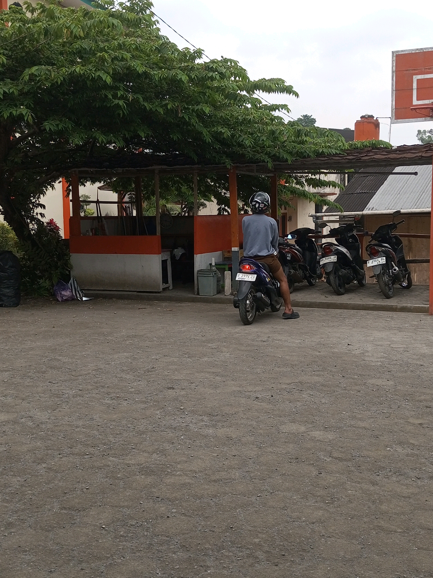 The image depicts an outdoor scene in Kalibeber, Indonesia, likely a parking area or a roadside. A person wearing a helmet and a gray long-sleeved shirt is seated on a blue scooter, facing away from the camera. The scooter is parked on a dirt or gravel surface. Behind the scooter, several other scooters are parked in a row. The background features a shaded structure with orange and white walls, possibly a small building or an outdoor shelter. A large tree with dense green foliage extends from the left side of the frame, casting shadows. A basketball hoop with an orange backboard is visible in the upper right corner of the image. The sky is overcast.