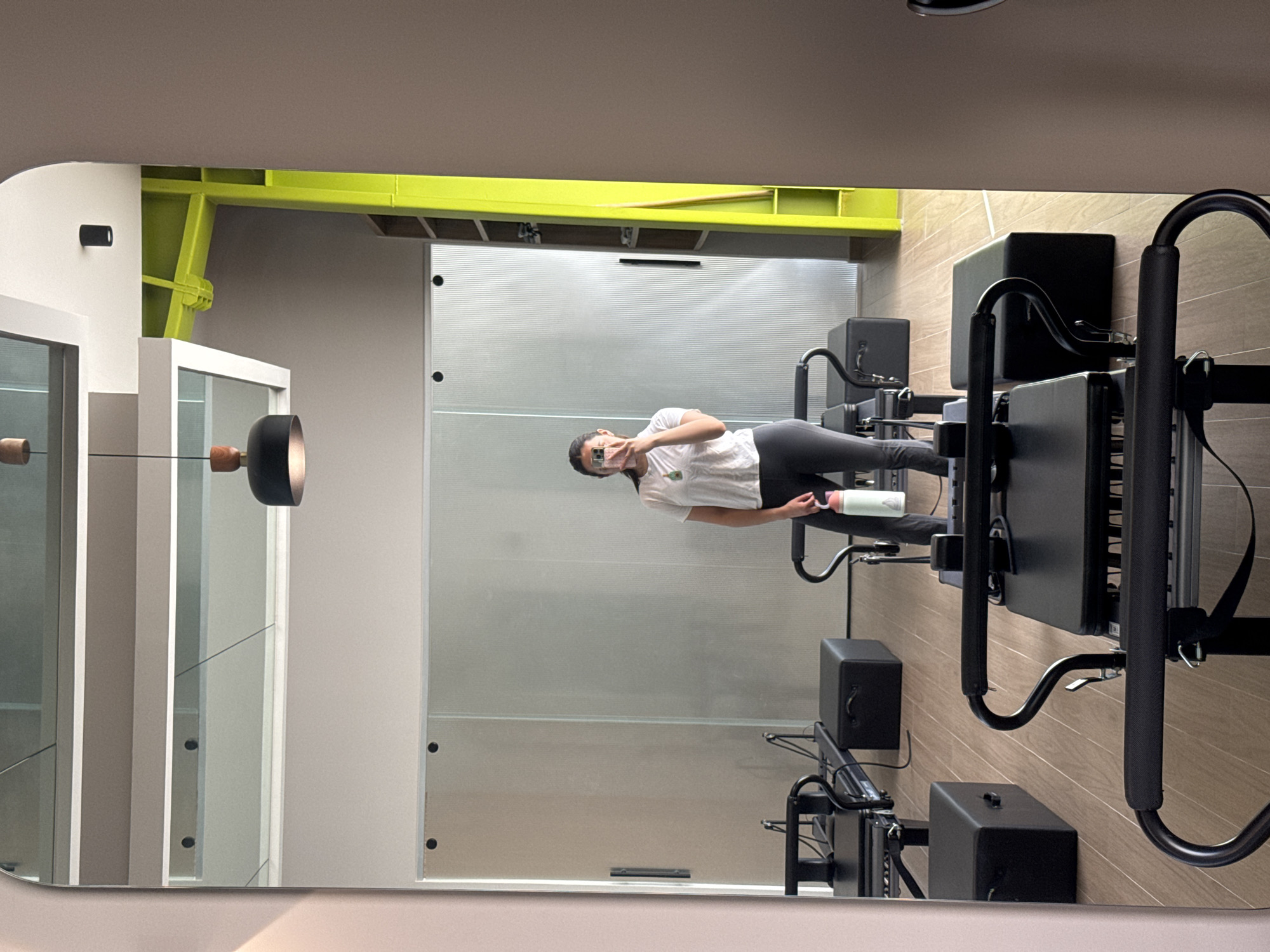 The image shows a woman reflected in a mirror inside a gym in San Bernardino Tlaxcalancingo, Mexico. She is standing next to a Pilates reformer machine. The room features a neutral color scheme with a light-colored ceiling, off-white walls, and a grey and tan striped background behind the reformer, possibly wallpaper or tiles. A yellow support structure is visible near the ceiling. Additional Pilates reformer machines are positioned next to the one by the woman. The mirror provides a partial view of a glass-paneled door or partition, and a modern pendant light fixture hanging from the ceiling. The woman is holding a water bottle.