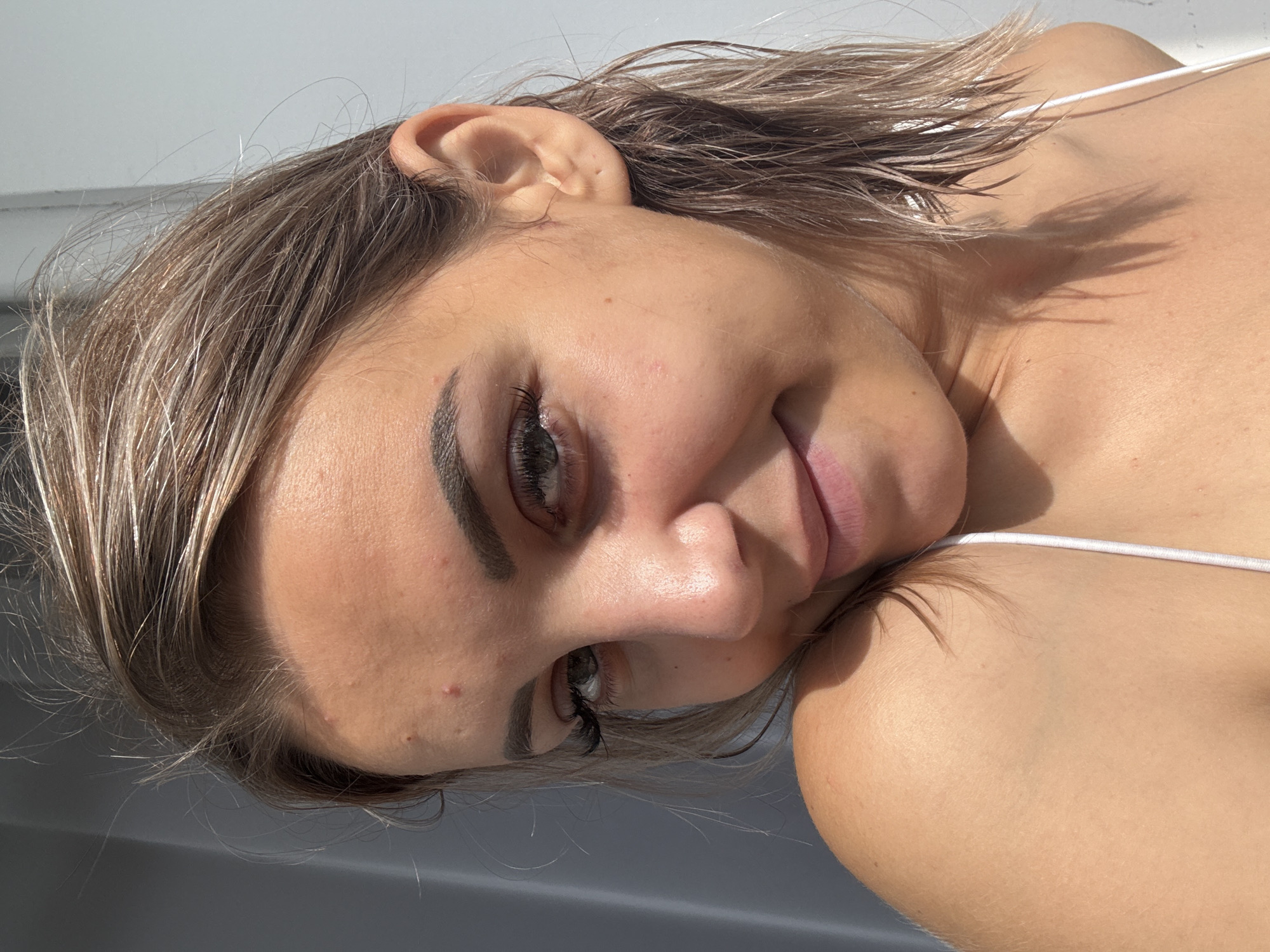 A young woman with light brown hair, styled in a wet, shoulder-length cut, is captured in a close-up, outdoor portrait. She has green eyes and is wearing a white bikini top. The lighting suggests it is a sunny day, with shadows cast on her shoulder and neck. Visible blemishes are present on her forehead. The background is a plain, light gray wall. The image does not contain any elements that indicate a specific location in Kholmsk, Russia.