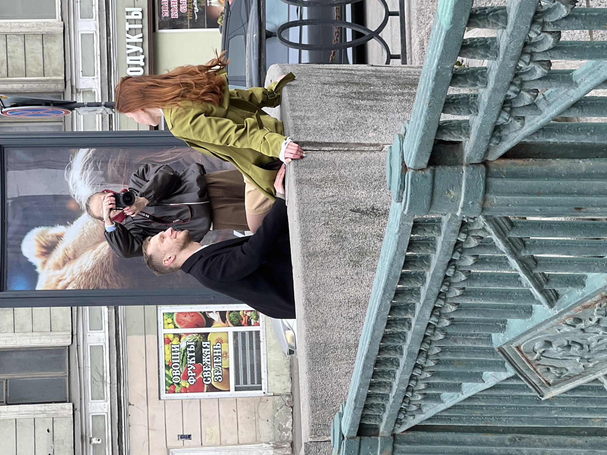A man and a woman pose on a bridge in Saint Petersburg, Russia. The woman, with long red hair and wearing a green trench coat, leans on the bridge parapet. A man in a black coat stands beside her, his hand touching the parapet near hers. A photographer captures the scene, standing on the street below with a camera. The backdrop features a storefront with windows displaying advertisements; one for "продукты" (products) and another for "ОВОЩИ ФРУКТЫ СВЕЖАЯ ЗЕЛЕНЬ" (vegetables, fruits, fresh greens). The bridge's metal railing is visible in the foreground. A car is parked near the store.