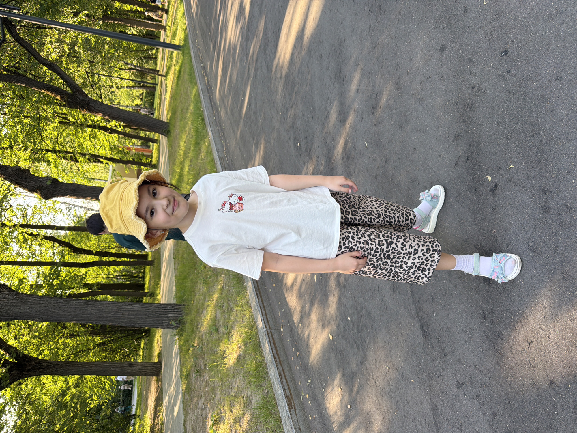 A young girl with a yellow bucket hat and a white t-shirt with a Hello Kitty graphic stands on a paved path in a park. She is wearing leopard-print capri pants, white socks, and light blue sandals. The park is filled with trees, and sunlight filters through the leaves, casting shadows on the ground. In the background, other people are visible walking. The scene appears to be a pleasant day in a city park, likely in Moscow, Russia, given the trees and style of the park.