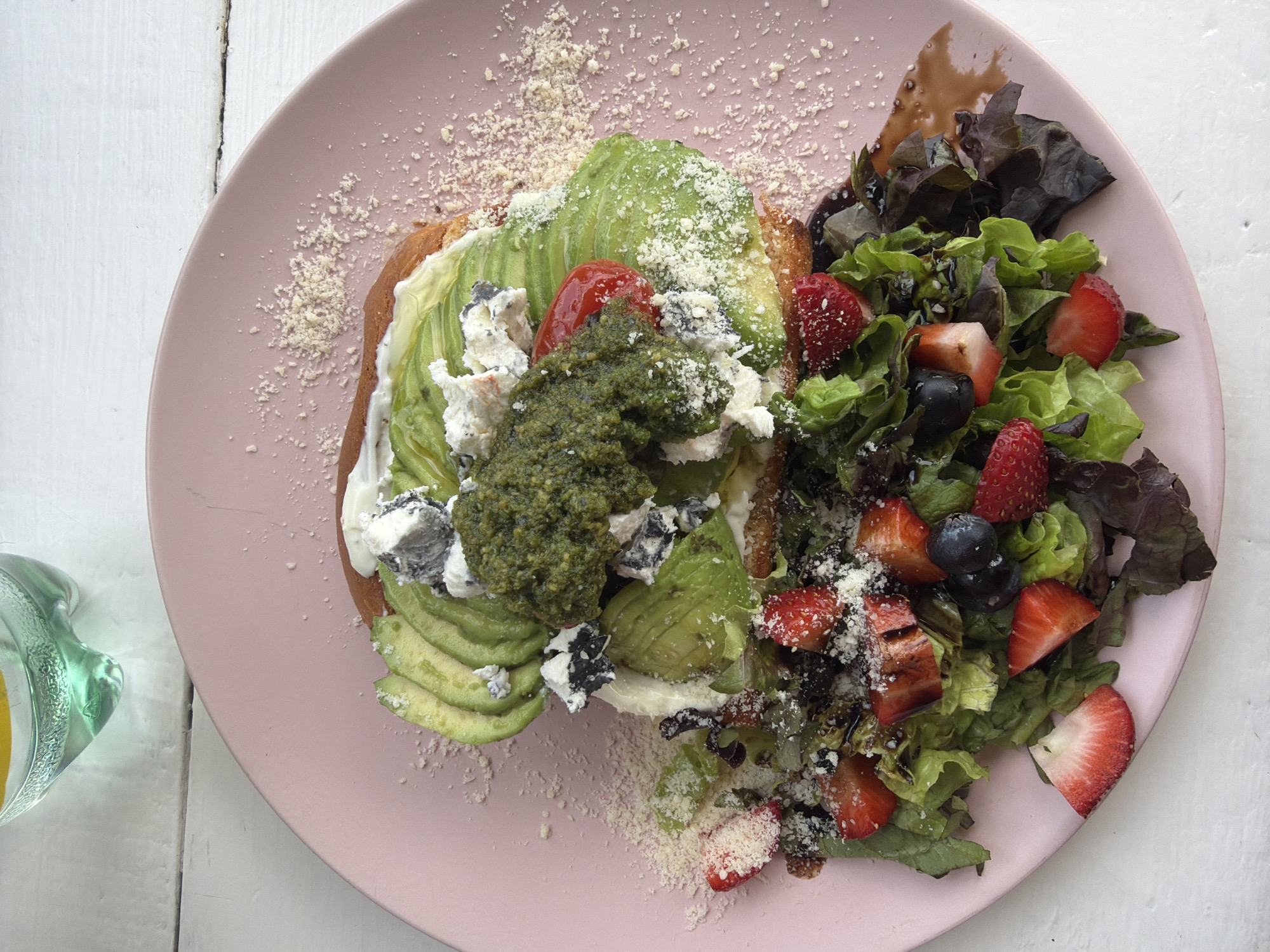 A top-down shot shows a pink plate featuring avocado toast and a side salad. The toast is topped with avocado slices, pesto, white cheese, and what appears to be parmesan cheese. The salad consists of lettuce, sliced strawberries, blueberries, and a dark dressing. The plate sits on a white wooden surface. A glass containing an orange liquid is visible in the lower-left corner of the frame. The location is Unknown, Unknown.