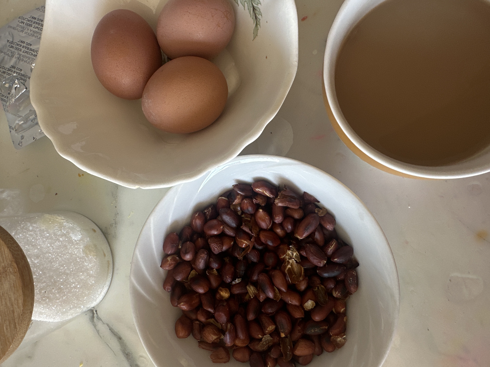 Here is a concise description of the image:
The image shows a tabletop with several food items. There's a bowl containing three brown eggs, a bowl filled with peanuts, and a mug filled with a light brown liquid, possibly coffee with milk. A jar with a wooden lid, potentially containing sugar, is also visible. The scene appears to be a simple breakfast or snack arrangement. Location information (Черёмушки, Russia) is not evident in the image itself.