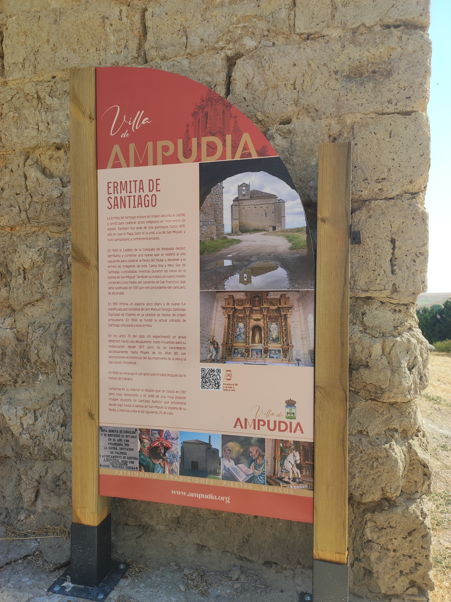 A vertical, weathered stone wall serves as the backdrop for a wooden-framed informational sign. The sign, made of laminated material, is attached to the wall by wooden posts. Its top section features a curvilinear red design with stylized white text that reads "Villa de AMPUDIA" and a coat of arms. Below this, a section with the heading "ERMITA DE SANTIAGO" provides historical information in Spanish.
The sign incorporates several photographic elements. One shows a small, stone building with a red tile roof and a bell tower, positioned under an arched opening within the sign's design. Another image displays an ornate, gilded altarpiece with multiple panels. A third section shows a collection of smaller images, one of which features a man in historical attire, and another depicting a white horse.
The lower portion of the sign has horizontal text and a website address. A QR code is also visible on the right side of the sign, likely intended for accessing further information. The environment surrounding the sign is dry and arid, with sparse vegetation and dry grass visible in the background, suggesting a warm, possibly Mediterranean climate. The stone wall itself is rough and shows signs of age and weathering.