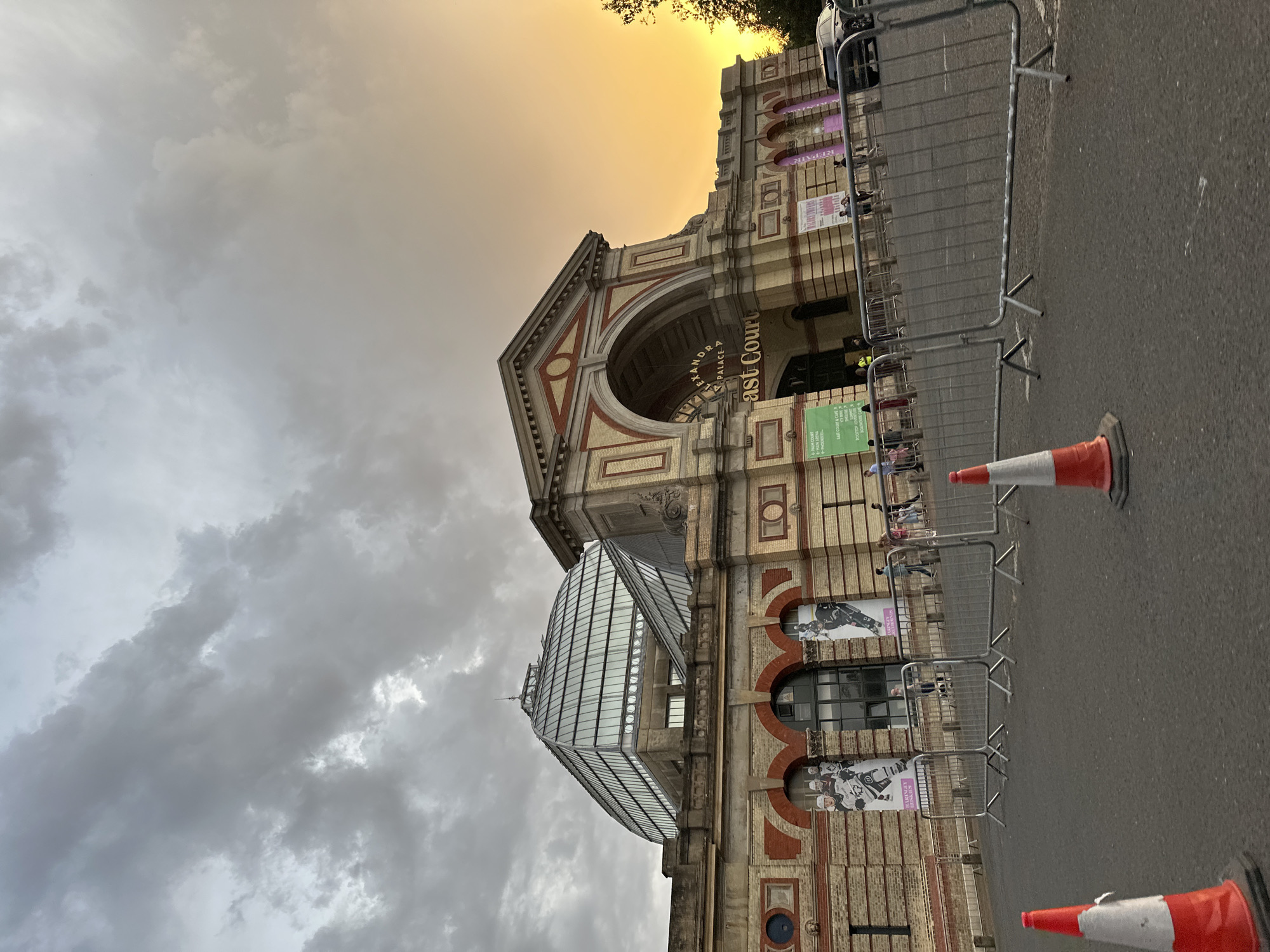 The image shows a street view of Alexandra Palace in London, England. The architecture of the building is visible, featuring brickwork, arched windows, and a glass structure extending from the side. A metal barrier runs along the pavement, with traffic cones placed at intervals. Posters are displayed on the building's facade, and people can be seen behind the barrier. The sky above is overcast, with a gradient of orange light towards the top of the building. The street surface is asphalt.
