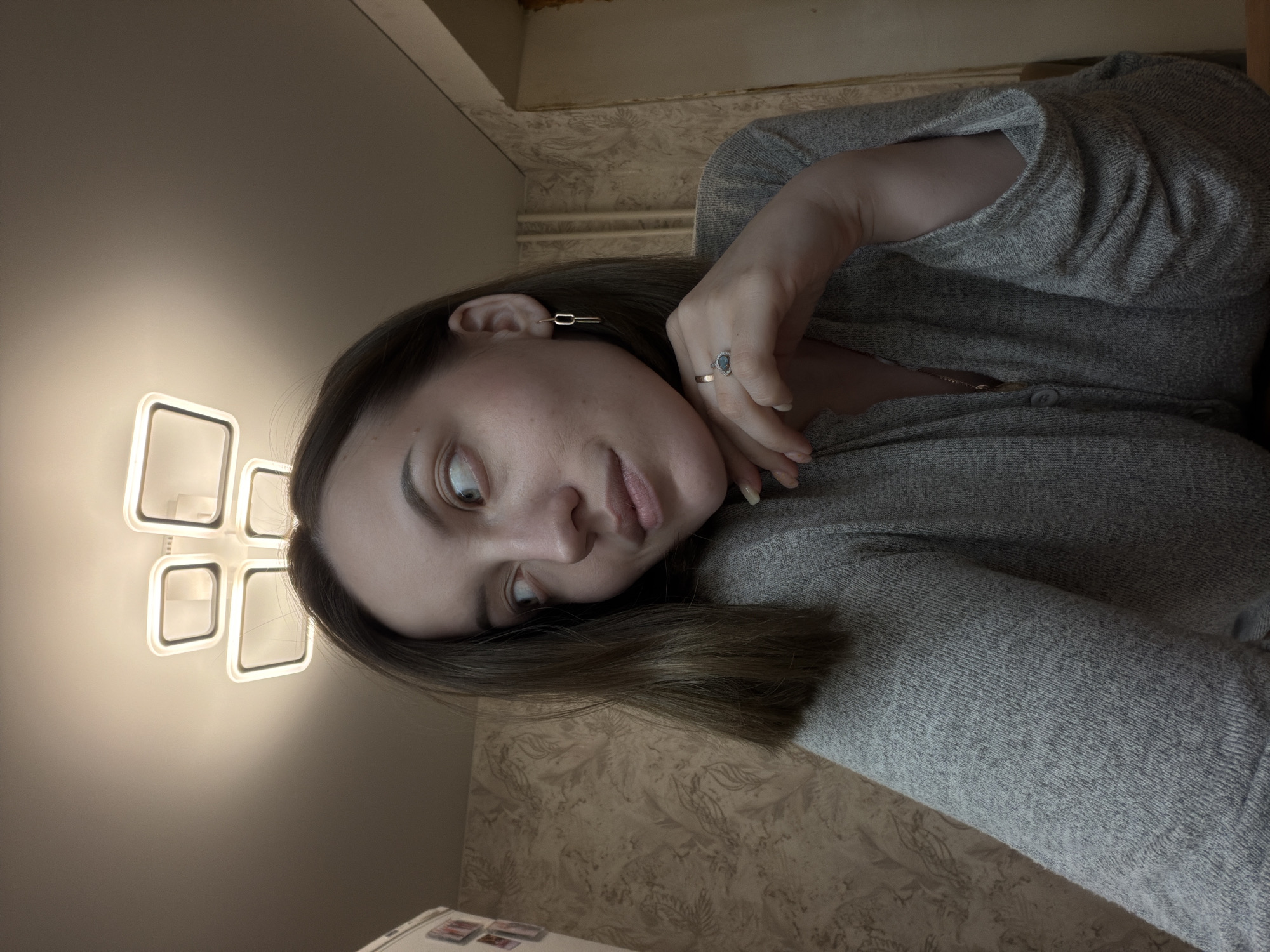 A woman with light skin and dark brown hair is facing the camera at an angle. She is wearing a gray knitted sweater. Her left hand is near her face, displaying a ring with a blue stone. She is in a room with wallpaper featuring a subtle leaf pattern. An ornate ceiling molding is visible at the top of the frame. A modern, square-shaped ceiling light fixture is positioned on the left side of the image. The location, Reshetnikovo, Russia, cannot be verified from the image.