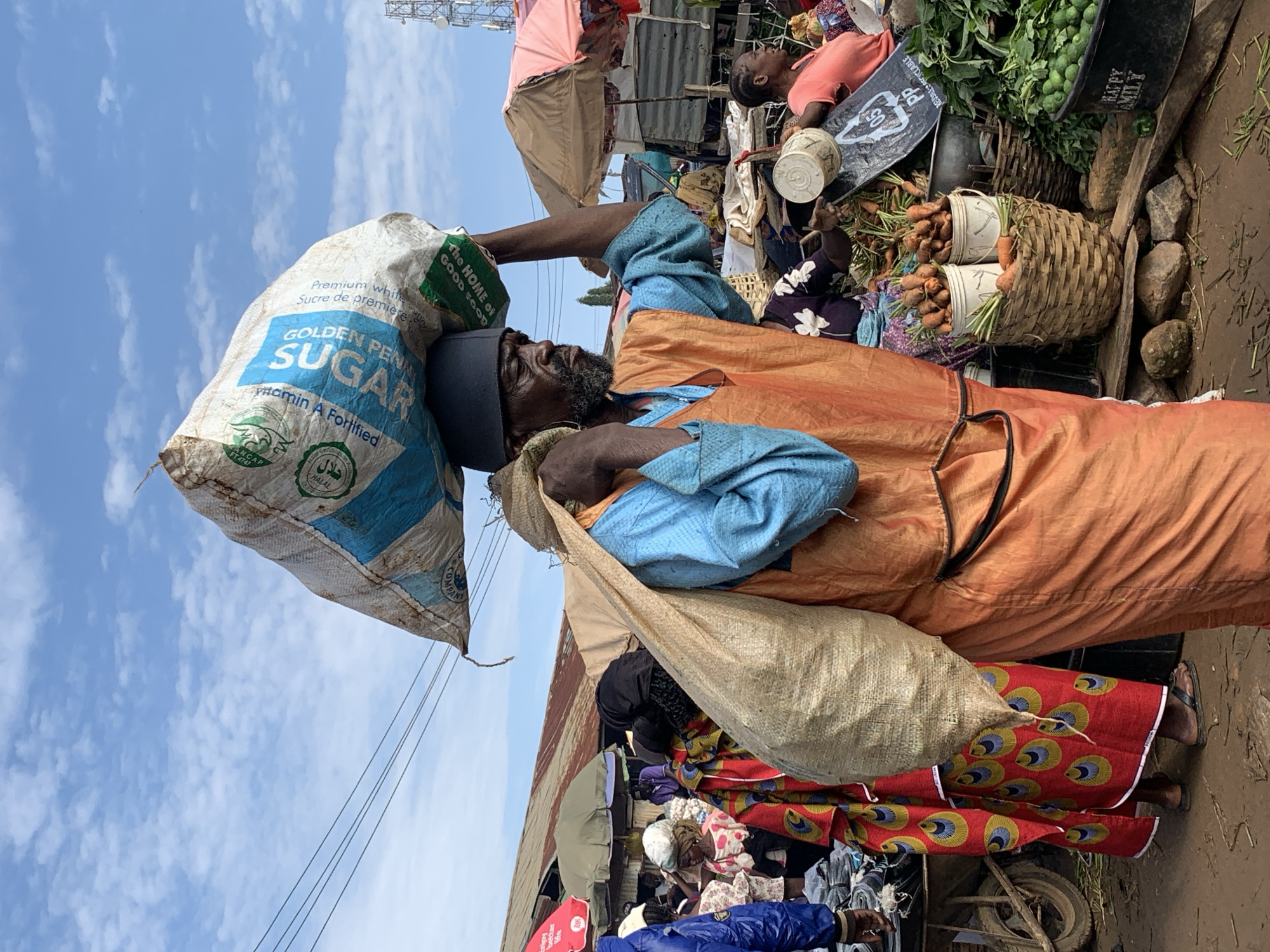 A man carries a large bag of sugar on his head while walking through a crowded outdoor market in Gura Top, Nigeria. He is also carrying another large sack over his shoulder. He is wearing a blue shirt and an orange vest. The market is filled with people, stalls, and produce, including carrots and leafy green vegetables. The ground is dirt and the sky is blue with scattered clouds.