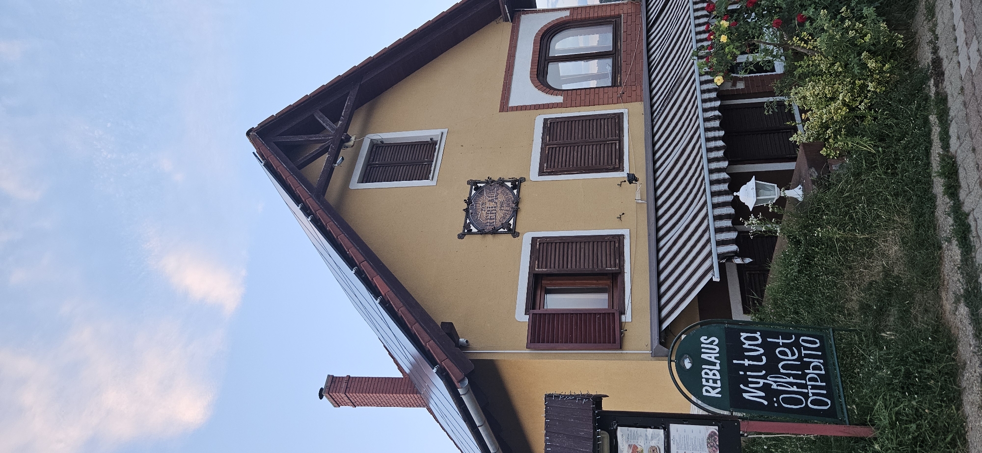 The image shows the exterior of a building in Hévíz, Hungary. The building is a two-story structure with a yellow facade and a red-tiled roof covered with solar panels. There are several windows with dark brown shutters. A sign in front of the building reads "REBLAUS Nyitva öffnet OTBblTO". A striped awning extends over the ground floor windows. In the foreground, there is lush green grass and a brick pathway. Red and yellow roses are visible in the lower right corner. The sky is a clear blue with a few wispy clouds. The overall scene depicts a business establishment, possibly a restaurant or guesthouse, in a residential or semi-rural setting.