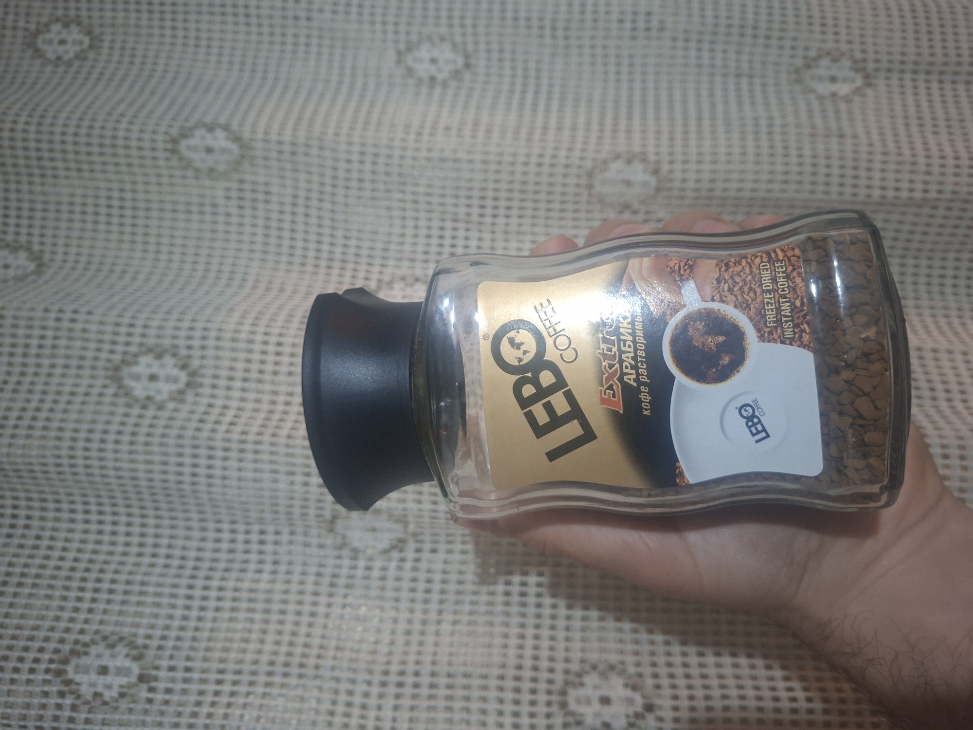 A hand holds a jar of "LEBO COFFEE" instant coffee against a patterned beige surface. The jar is clear, allowing the coffee granules inside to be visible. The label is gold and black with an image of a coffee cup. The jar has a black lid. The location is unknown.