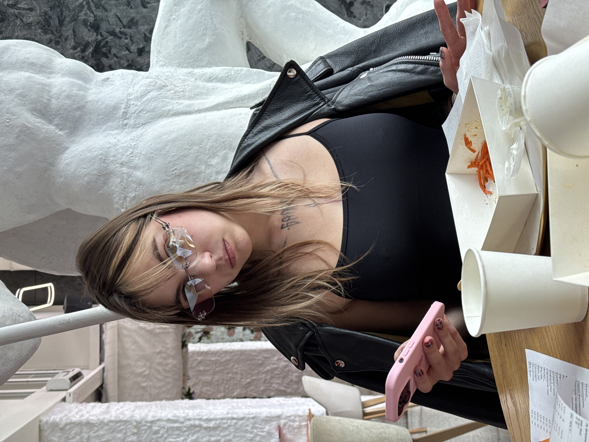 A young woman with blonde and brown hair, wearing rimless heart-shaped glasses, a black tank top, and a black leather jacket, sits at a wooden table. She is holding a pink smartphone in her left hand and gesturing with her right hand. In front of her are a white paper cup, a white takeout box with shredded carrots, a small container of sauce, and a napkin. There is also a receipt on the table. The background shows a textured white pillar and a large white abstract sculpture. The overall setting appears to be a cafe or restaurant.