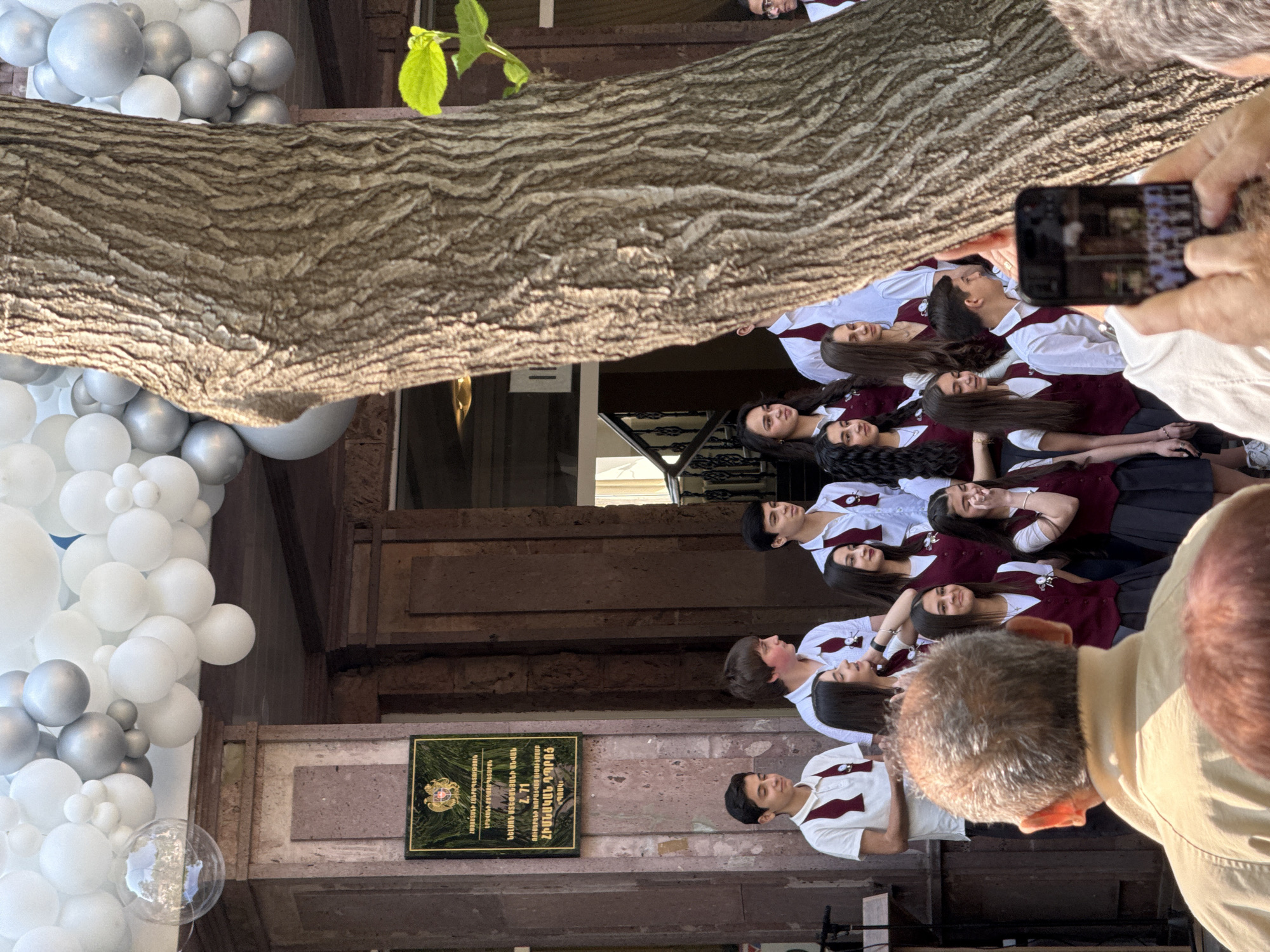 A group of about a dozen students in school uniforms are posing for a photo in front of a building, likely in Yerevan, Armenia. The students are wearing white shirts, maroon vests, and dark skirts or pants. A person is taking a photo of the group with a smartphone. There are white and silver balloons to the left of the building's entrance. A plaque with Armenian text is on the building's facade. A large tree branch obscures part of the scene.