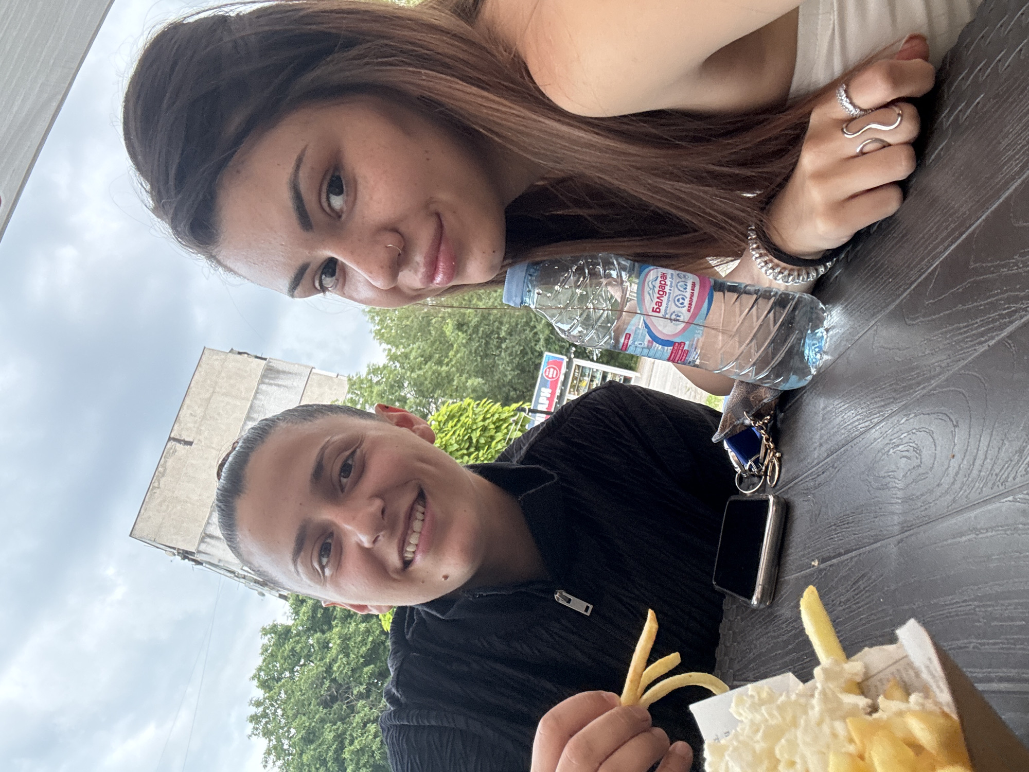 Two young individuals are seated at a dark wood-grain patterned outdoor table, looking directly at the camera under an overcast sky. The person on the left, with dark hair pulled back into a bun, wears a black textured half-zip top and holds a cluster of French fries in their right hand. They exhibit a mole near the right corner of their mouth. On the right, a person with long brown hair and freckles wears a white strapless top. This individual has a silver stud nose piercing and a silver ring lip piercing, and holds a clear plastic bottle of "Балдаран" (Baldaran) brand bottled spring water, featuring Cyrillic text. Their right hand displays a wavy silver ring on the middle finger and a silver band with stones on the ring finger. A dark smartphone and a set of keys with a fob are visible on the table between them. A paper cone of French fries, topped with a substantial amount of a white, fluffy substance, is situated to the left. The background consists of lush green trees and an older, light-colored concrete multi-story building showing signs of weathering. A distant partial sign with red Cyrillic letters "АРИ" is visible, confirming the location as Sofia, Bulgaria, due to the combination of the Bulgarian water brand and Cyrillic script.