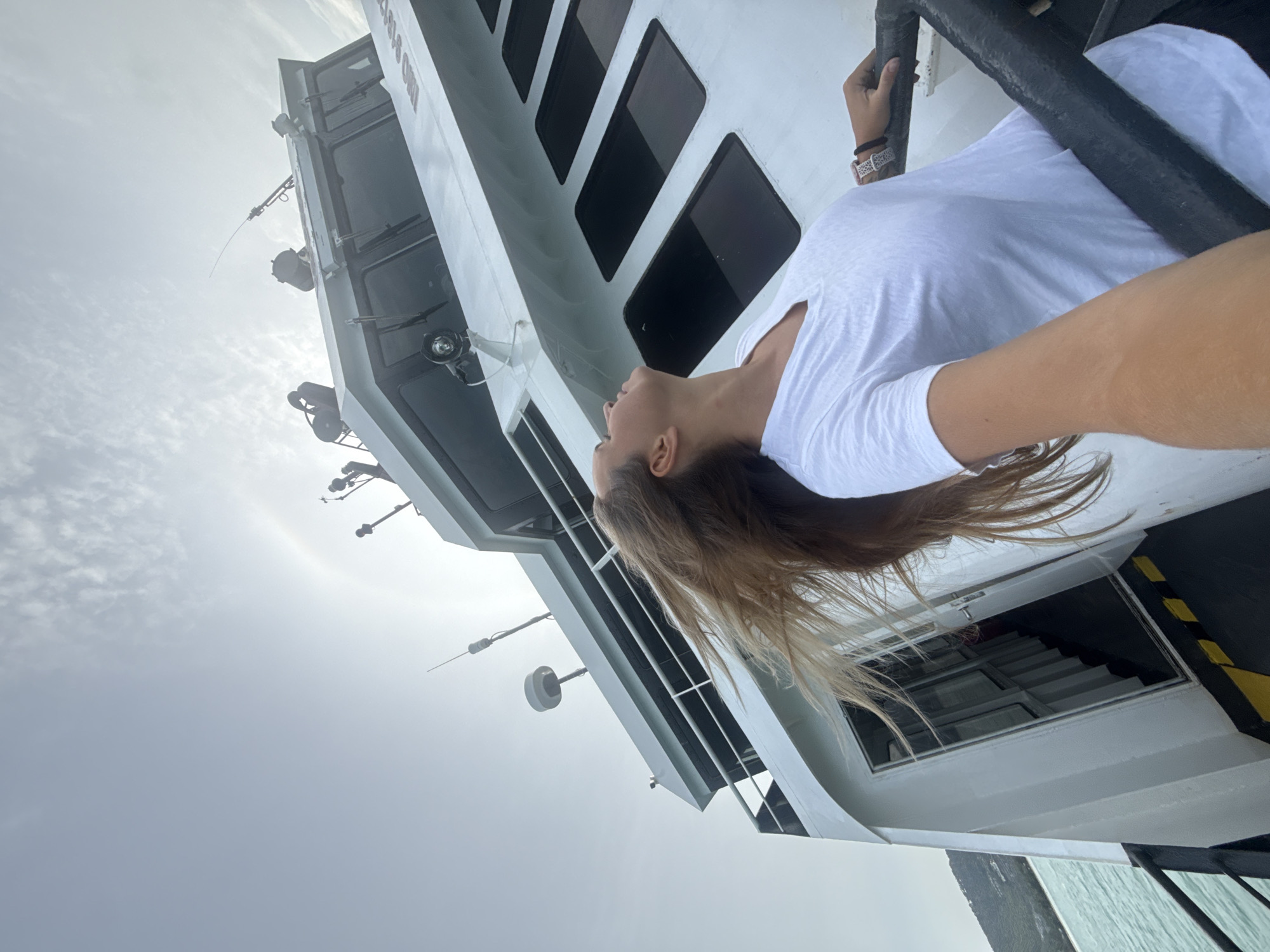 A young woman with long, blonde hair is taking a selfie while standing on the deck of a ferry. She is wearing a white t-shirt and looking up at the sky. The ferry is white with black railings. The sky is overcast with a visible sun halo. The background shows a body of water and a green, mountainous island. The location is Ko Samui District, Thailand.