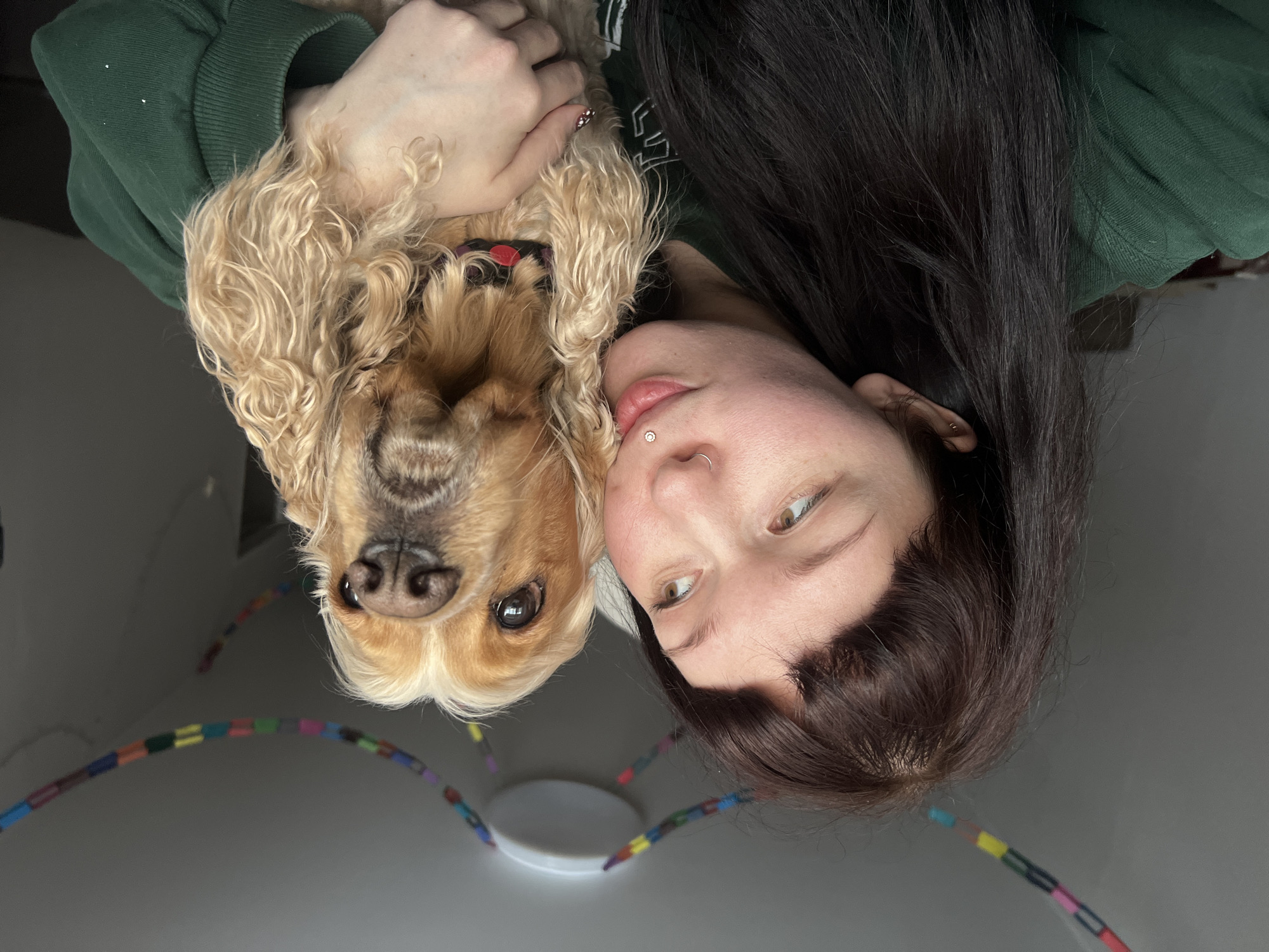The scene depicts an indoor, close-up portrait of an individual and a dog in Chaikovsky, Russia. A young adult female with long, dark brown hair and bangs is positioned on the left side of the frame. She has fair skin, light brown eyes looking towards the right, and two facial piercings: a silver stud above her upper lip and a silver hoop in her right nostril. Her mouth is slightly open, appearing to gently touch the dog's head. She is wearing a dark green long-sleeved garment. Her right hand is visible, holding the dog, with some fingernails exhibiting a red and white checkered pattern. The dog, identified as a Cocker Spaniel or similar breed, possesses light brown and white wavy fur, dark eyes, and a dark nose. It is positioned to the right of the individual, looking directly at the camera, and wears a collar with red and black components. The background consists of a plain, light-colored wall and ceiling. A circular white ceiling light fixture is visible centrally on the ceiling, from which a colorful garland, composed of segmented links in various hues, is strung in multiple arcs across the upper portion of the frame.