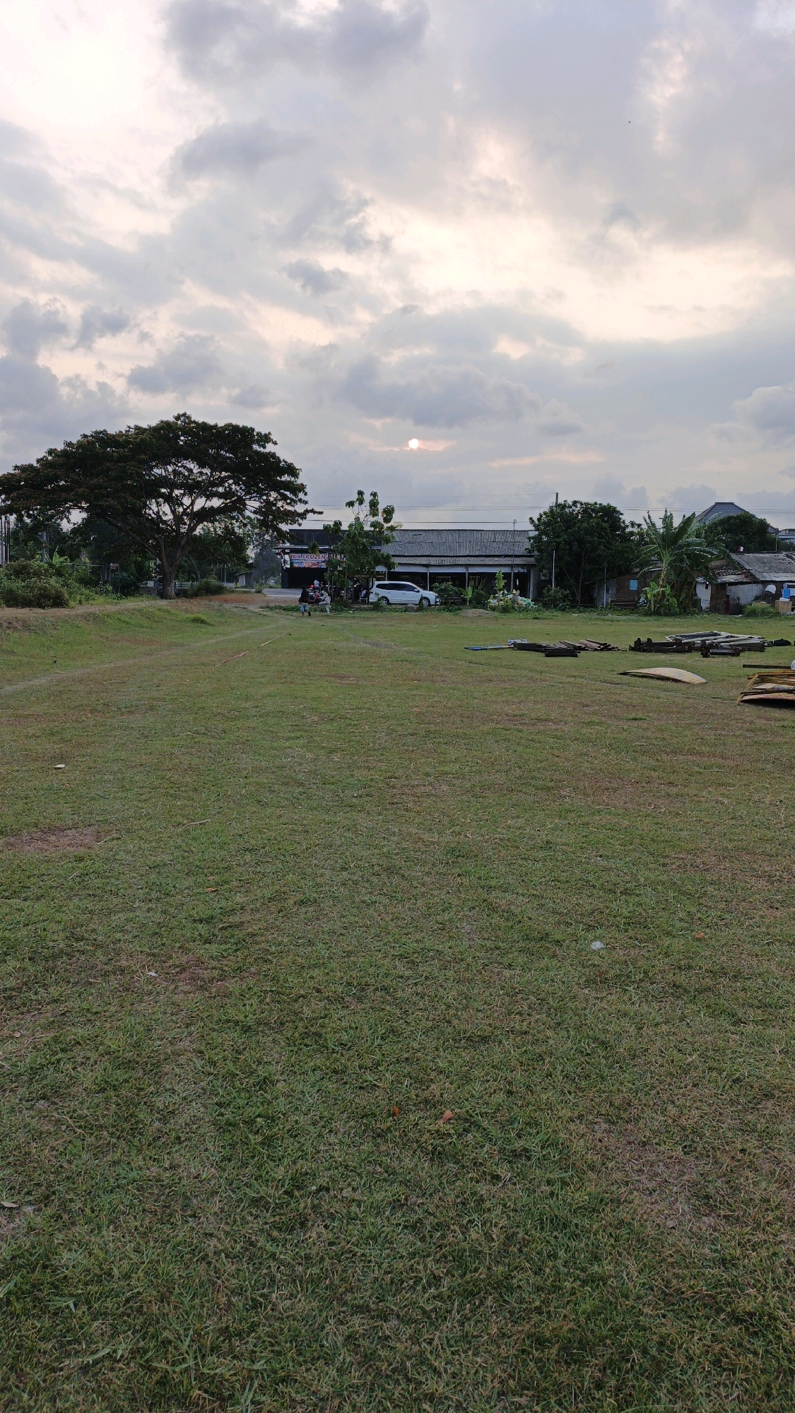 An outdoor, low-angle, vertical, full shot shows a large, mostly empty grassy field in Boyolangu, Indonesia, under a cloudy sky with the sun partially obscured by clouds. The field is a muted green with some dry patches and is mostly free of debris, with a small pile of assorted wooden planks and beams stacked on the right side of the frame. In the middle ground, a paved road runs horizontally across the scene, bordered by buildings, trees, and vegetation. Several people are visible on or near the road. A white SUV is parked on the side of the road. The background is filled with a mixture of residential and commercial buildings, surrounded by lush green trees and vegetation. The sky is overcast with scattered clouds that are lighter in color due to the sun's presence behind them. The overall impression is of a quiet, rural area on a cloudy day.