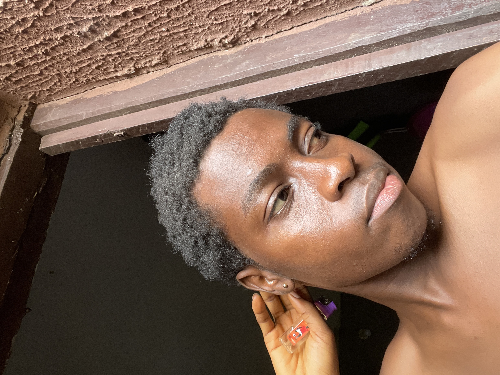 A person with dark skin and short, dark curly hair is posed in front of a window, looking to the side. They are holding a small, transparent object with a cartoon design on it near their ear. The window frame is brown, and above it is a textured ceiling. The location is likely Nkpor, Nigeria. The person's neck and shoulder are visible, and they appear to be shirtless. There is a dark, undefined background visible through the window.