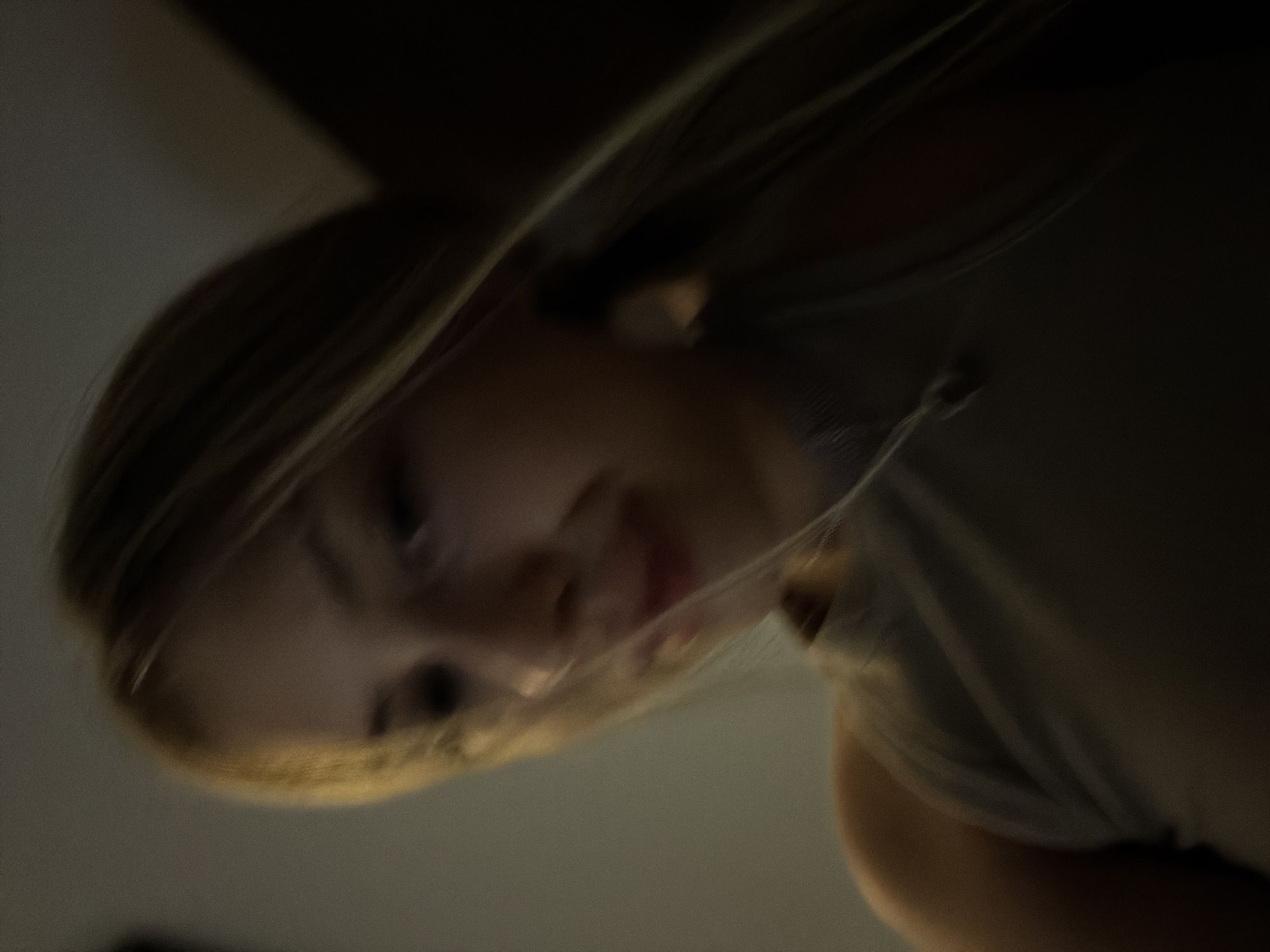 The image shows a close-up, blurry shot of a person, likely a young woman, lying down. Her blonde hair is visible, as is part of her face, including her nose and mouth. She appears to be wearing a light-colored shirt or top. The lighting is dim, casting a shadowy effect on the scene. The background is indistinct, but suggests an indoor setting with a plain wall. The image is not specific to any location in Đà Nẵng, Vietnam, or any identifiable city.
