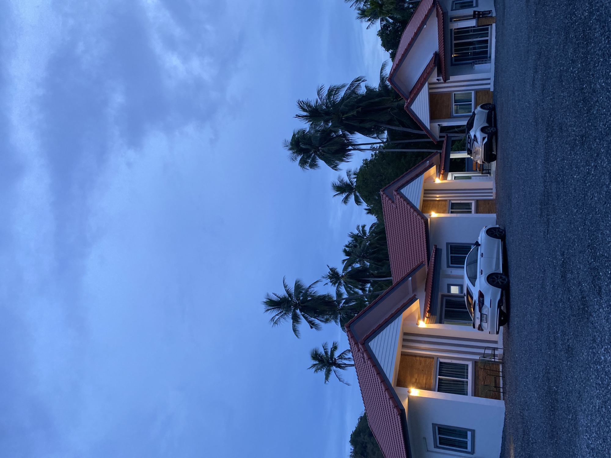 The image shows a row of single-story buildings with red tile roofs and white facades in Baan Harn, Thailand. Palm trees are interspersed between the buildings. A white car is parked in front of one of the units. The sky is a dusky blue, indicating early evening. The ground appears to be a paved surface. No people are visible.