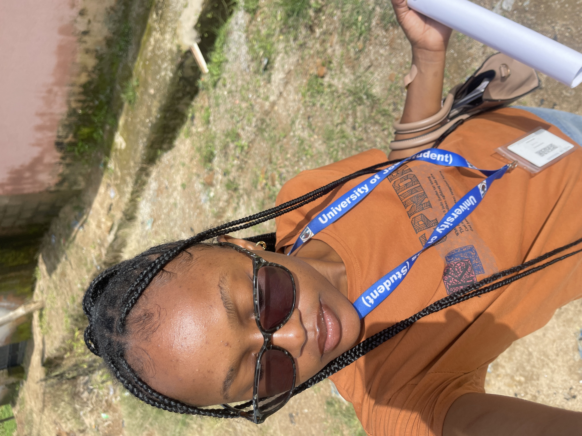 A young Black woman with braided hair and sunglasses is standing outdoors, holding a rolled-up document and a light brown handbag. She is wearing an orange t-shirt with a lanyard around her neck that reads "University of Jos (Student)". The background suggests an outdoor urban or semi-urban environment, possibly near a building with a pinkish-red wall. The ground appears to be a mixture of dirt, gravel, and some vegetation. The scene is likely in Jos, Nigeria, given the university's affiliation.