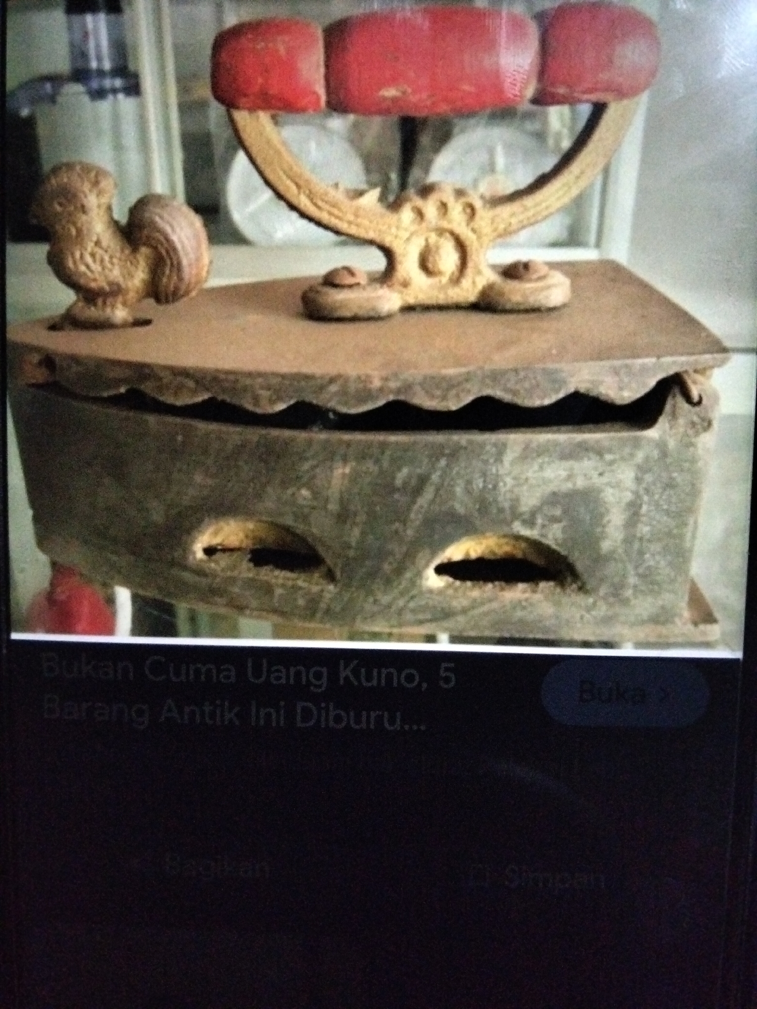 This is a close-up, eye-level, outdoor shot of an antique charcoal iron displayed on a weathered wooden surface. The iron is made of dark metal and has a decorative rooster figure perched on its lid, which is embellished with red ceramic handles. The iron has a wavy edge and two oval-shaped openings on its side. The background is blurred and suggests a display area, possibly a market stall or antique shop, with glimpses of other items and a reflective surface. A portion of a digital screen with Indonesian text and icons is visible at the bottom, indicating the image might be a screenshot or viewed on a device. There is no visible presence of people, and the environment appears to be a commercial or exhibition setting, not specifically within Kualaraja, Indonesia.