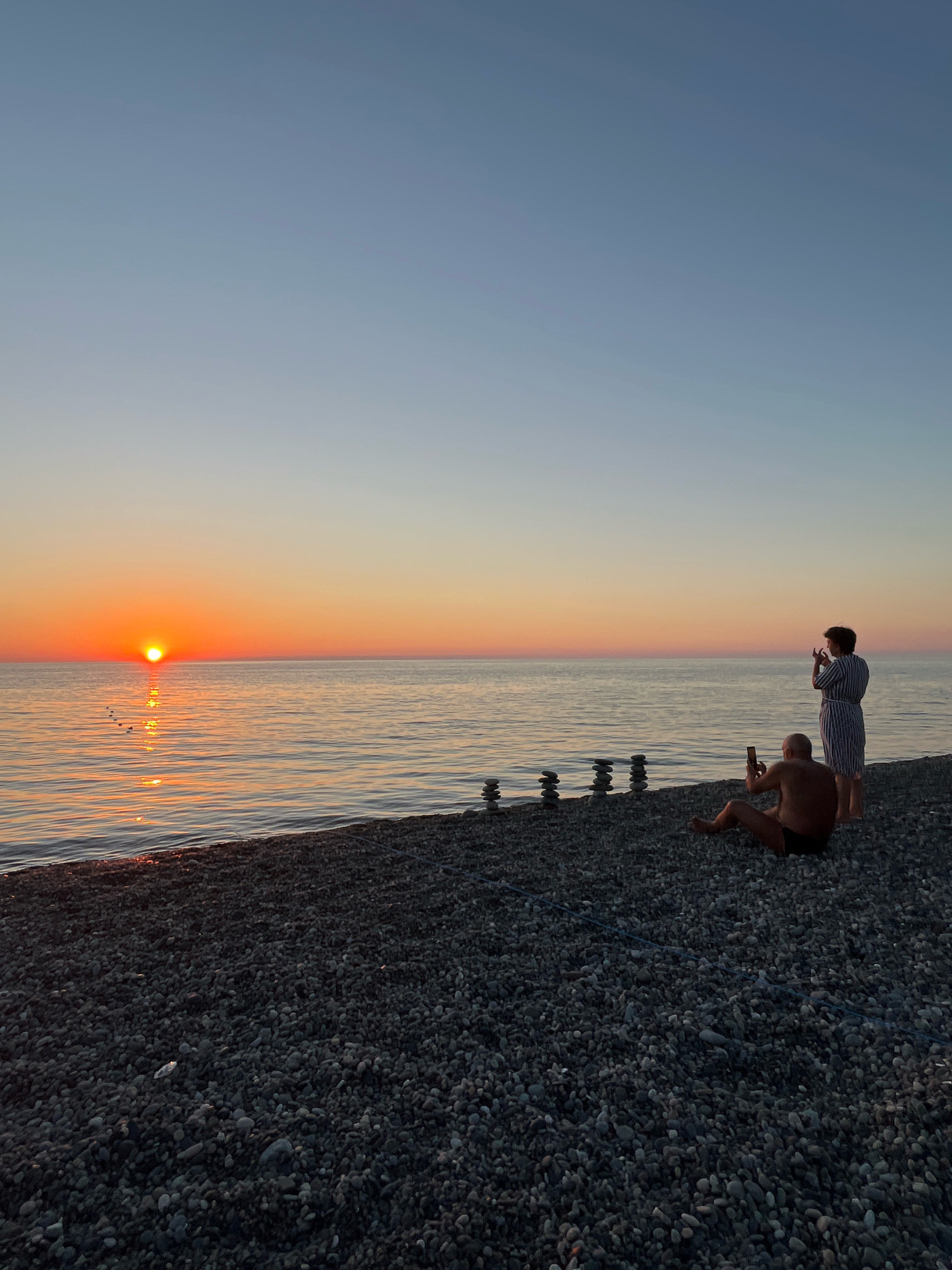 This is a vertical, eye-level, medium shot of a sunset over the Black Sea on the pebble beach of Kobuleti, Georgia. The sun, a bright orange disc, is low on the horizon, casting a shimmering reflection on the calm water. The sky above is a gradient of soft blues and oranges, transitioning to a pale yellow near the horizon.
In the foreground, on the dark grey pebble beach, are two people. A man, wearing dark swim trunks, is sitting on his left side, facing the sea and holding a smartphone in his right hand, appearing to take a photo or video. To his right and slightly behind him, a woman with short dark hair, wearing a vertically striped blue and white dress, is standing and also holding a smartphone in her left hand, seemingly capturing the sunset.
Between the two people and closer to the water's edge are several stacks of balanced stones, arranged in a somewhat artistic fashion. The water is tranquil, with small ripples reflecting the warm hues of the sunset. The beach is covered in smooth, dark grey pebbles of various sizes. The overall scene evokes a sense of peaceful observation and appreciation for the natural beauty of the coastal sunset.