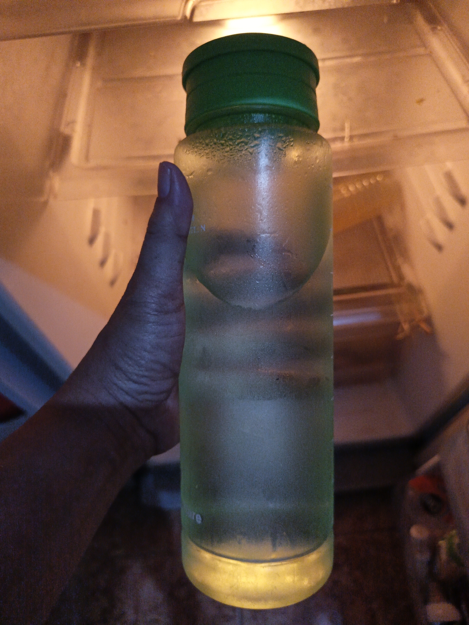 A hand is holding a frosted green-lidded water bottle inside a refrigerator. The bottle is filled with a light-colored liquid and has condensation on its exterior. The interior of the refrigerator is dimly lit, with shelves and compartments visible. The overall scene suggests a cool environment, likely in Mojayan, Indonesia, where the water bottle is being kept chilled.