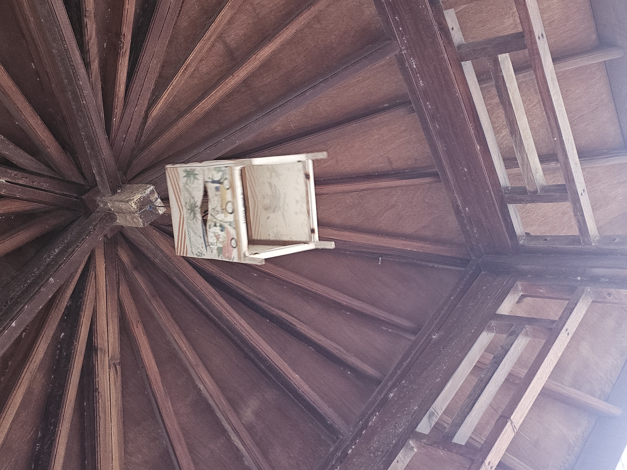 An interior view looking up at the wooden ceiling structure of a gazebo or pavilion in Dalegan, Indonesia. The ceiling is made of dark wood planks radiating outwards from a central point. Suspended from the ceiling is a small, multi-tiered wooden structure, possibly a birdhouse or decorative element, adorned with a colorful painted scene depicting palm trees and people. The lower section of the structure shows framing and wooden planks, suggesting it is part of a larger building. The lighting is soft, casting gentle shadows on the wooden surfaces.