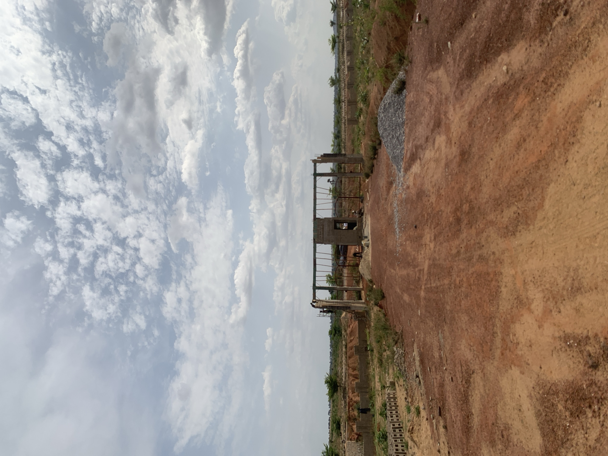 A photograph captures a construction site in Kujama, Nigeria. The sky, filled with white, layered clouds, occupies approximately half the frame. Below, an expanse of reddish-brown soil dominates, with a partially constructed metal gate structure positioned centrally. The gate is not complete, and construction is ongoing. Scattered grey gravel lies to the right of the gate, amidst the reddish-brown earth. In the distance, vegetation and other structures, possibly dwellings, are visible, suggesting a developing urban or peri-urban landscape. No people are immediately visible within the frame.