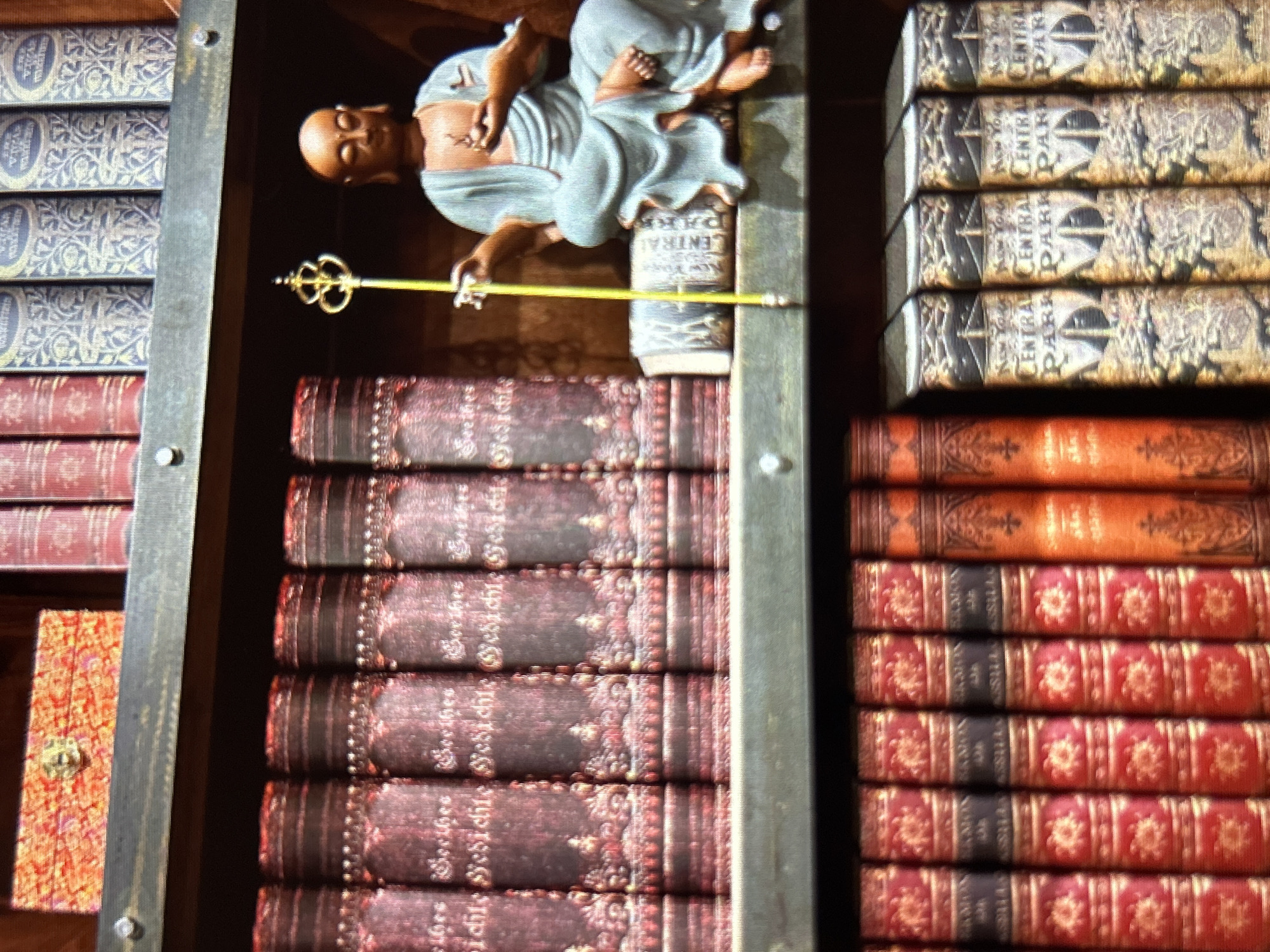 This media file is a close-up, tilted shot of a shelf filled with books and a decorative statue. The statue depicts a bald figure in flowing blue robes, reclining on a column and holding a golden scepter. The books are varied in appearance: some are bound in red with intricate gold patterns on their spines, while others have gold and dark blue patterned covers with visible text. The setting appears to be indoors, possibly a library or a study, with the lighting suggesting an artificial source. The overall aesthetic is one of antique and decorative collections. The visible text on some book spines includes "CENTRAL PARK," "NEW YORK," and what appears to be "Gesichte" in German.