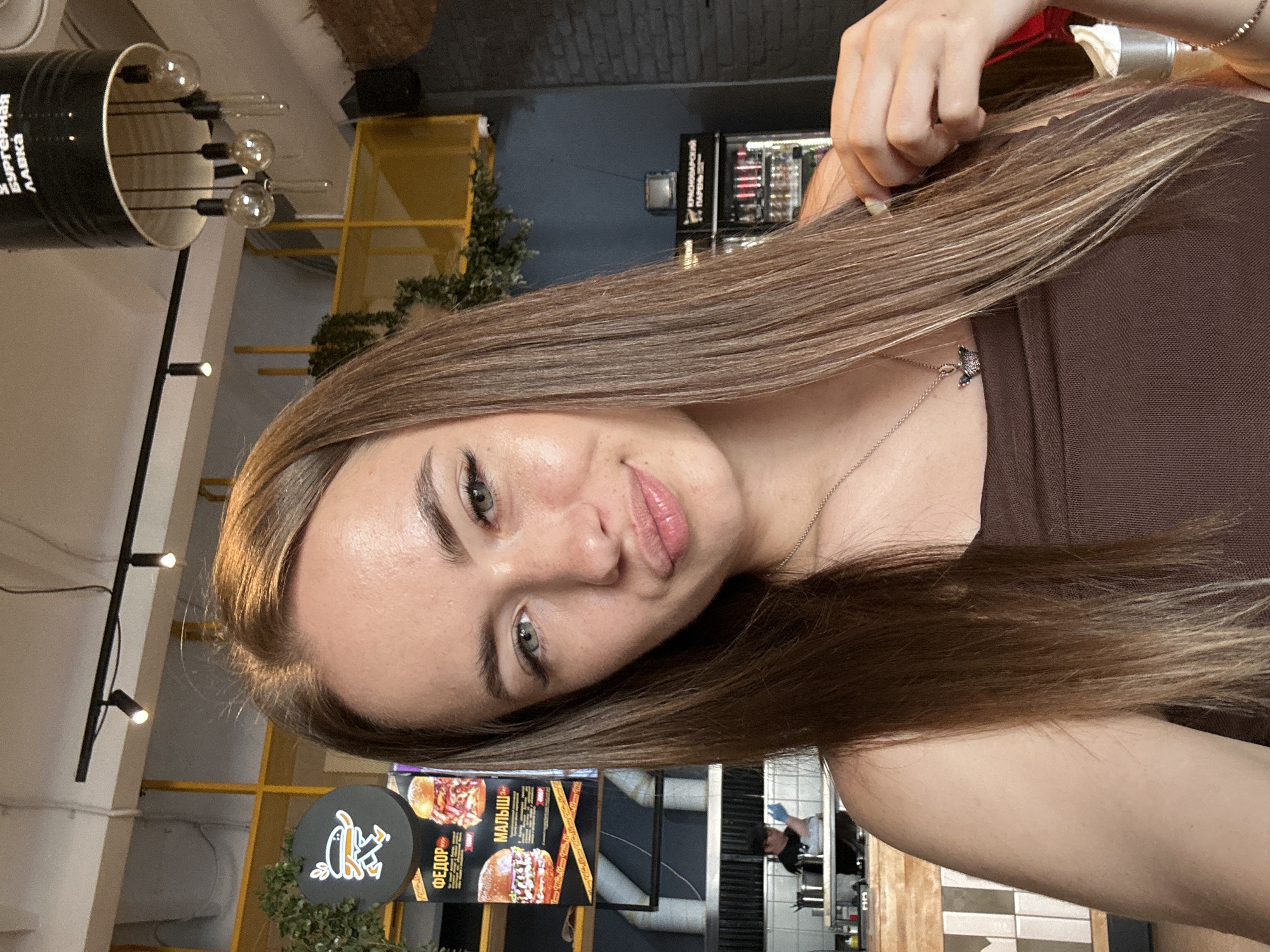 A young woman with long, straight brown hair and green eyes is taking a selfie in a restaurant. She is wearing a brown top and a silver necklace with a small pendant. The background shows a yellow shelf unit with greenery, a menu board with pictures of burgers, and a glimpse of a kitchen area where an employee is visible. The restaurant appears to be casual, possibly a burger joint, with industrial-style lighting and brick accents.