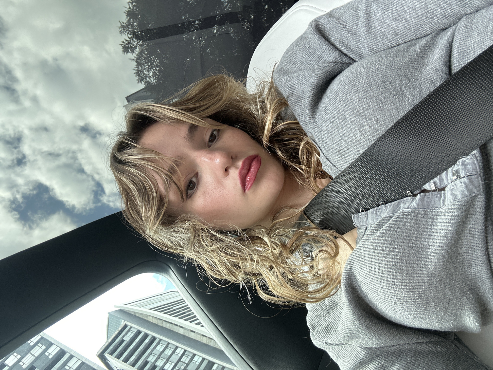A young woman with blonde, wavy hair and full lips with red lipstick is pictured in the driver's seat of a vehicle. She is wearing a grey, ribbed top and a black seatbelt is across her chest. The background shows a city skyline with modern buildings, suggesting an urban environment. The sky above is partly cloudy. This image was taken in San Bernardino Tlaxcalancingo, Mexico.