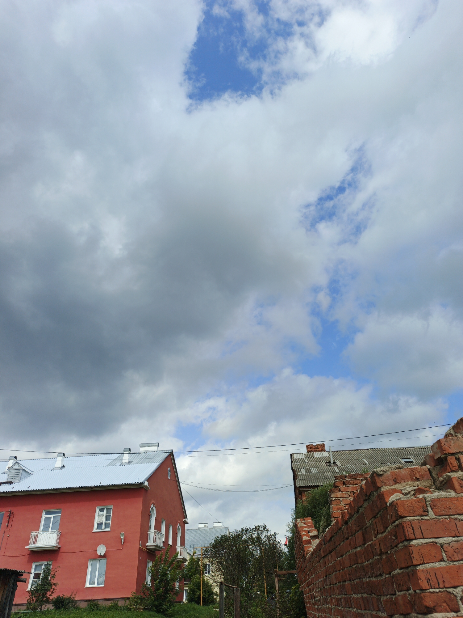 In Medvedyovka, Russia, an image captures a cloudy sky with patches of blue over a residential area. A bright red, multi-story building with a silver roof and white-trimmed windows is on the left. Balconies with white railings jut out from the red building. To the right, a damaged red brick wall with a grey, corrugated roof building behind it dominates the foreground. Trees and green shrubbery are visible along the bottom of the frame. Power lines are strung across the scene.