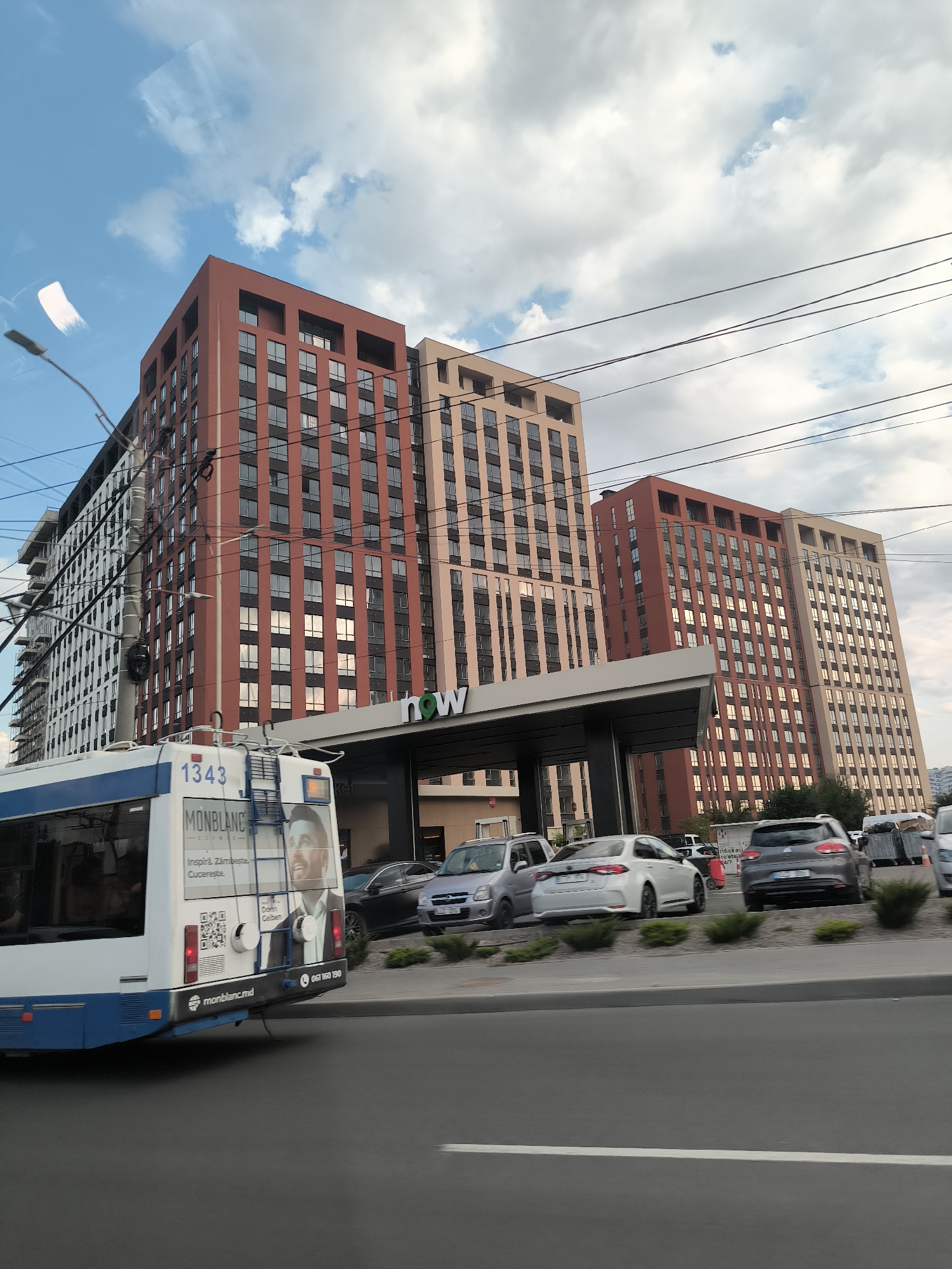 The image depicts a street scene in Chișinău, Moldova, featuring a modern apartment complex with distinct red and beige facades, and a gas station canopy labeled "now" in green lettering. A white and blue trolleybus is partially visible on the left, displaying the number "1343" and advertising "MONBLANC" with a man's portrait. Several cars are parked in front of the buildings, including a white sedan, a dark grey hatchback, and a silver hatchback. The environment includes a sidewalk with manicured bushes, overhead power lines, and a cloudy sky. The perspective suggests the photo was taken from a moving vehicle.