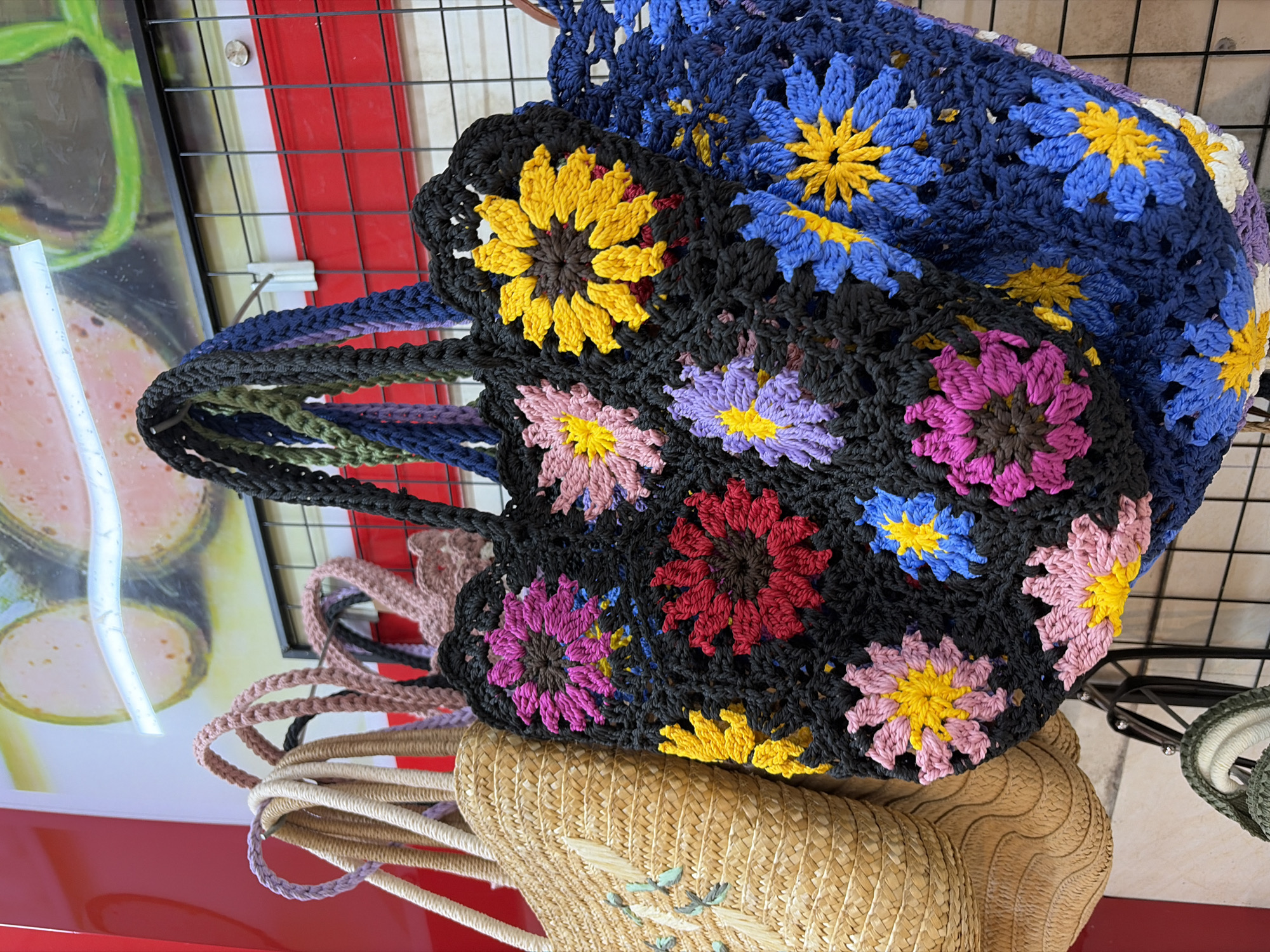 The image displays a close-up view of several woven bags hanging on a display rack, likely in a market or shop in Da Nang, Vietnam. The bags are diverse in material and design. On the left, a straw tote bag with embroidered light blue floral designs is visible. To its right, a black crocheted tote bag is prominently featured, adorned with colorful crocheted flowers in shades of pink, red, yellow, and purple. Another crocheted bag, primarily in shades of blue and yellow with some white and purple accents, is partially visible to the right of the black bag. The background includes a red wall and a framed image with depictions of what appears to be sliced meat. The display rack is a black grid, and the items are illuminated by overhead lighting.