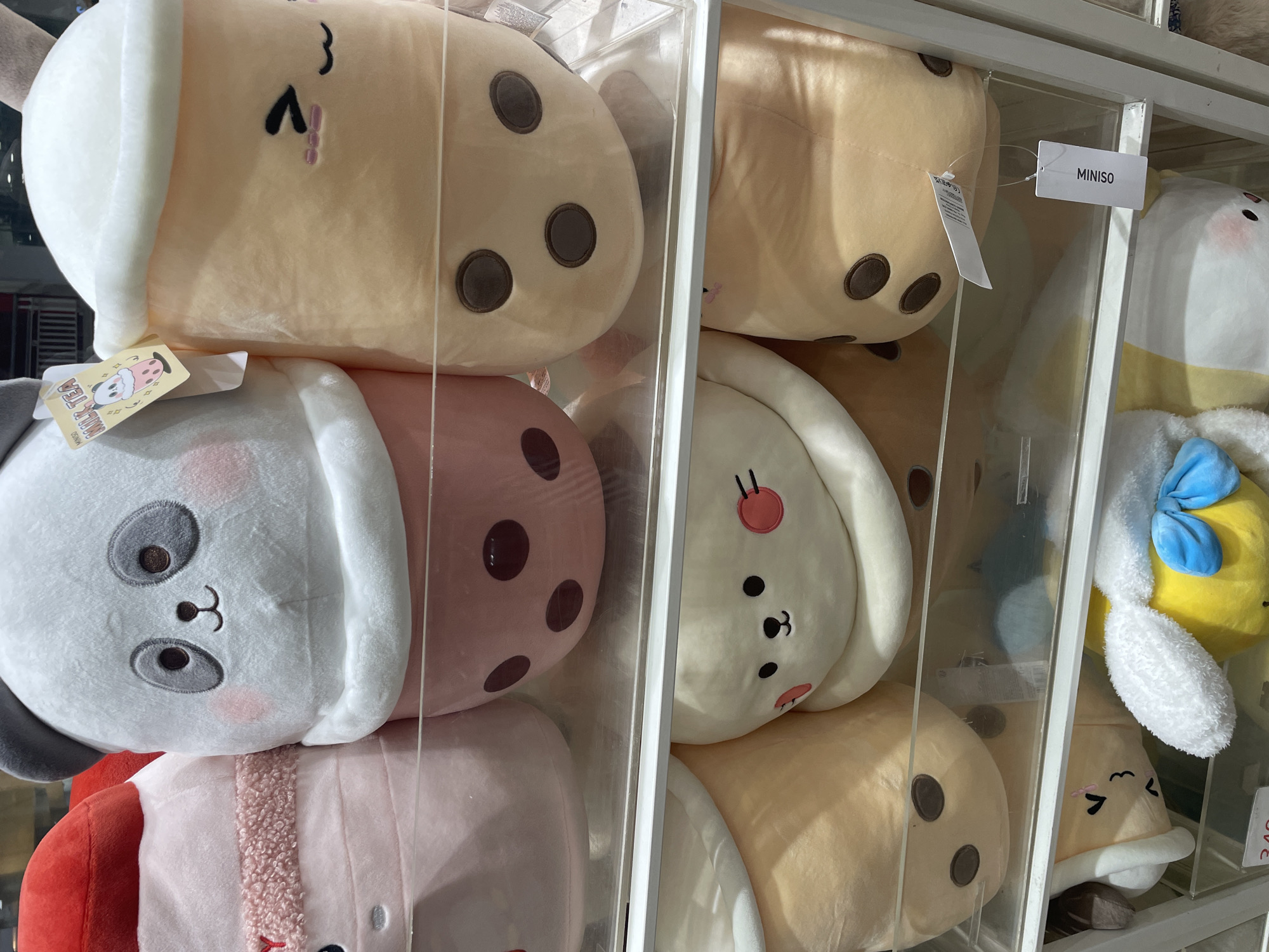 The image shows a shelf display of large plush toys, possibly at a Miniso store in Davao City, Philippines. The toys are arranged in clear plastic compartments. The top shelf features a light pink plush toy with a fuzzy white trim on the left, and a light grey panda-themed plush toy with pink cheeks and dark grey ears. Next to the panda plush is a large, light orange plush toy shaped like a cup of milk tea, with a white frothy top and a cute smiling face with closed eyes and pink blush marks. The milk tea cup has dark brown "boba" spots. The middle shelf displays two plush toys. The one on the left is a light orange plush toy with three dark brown boba spots. The one on the right is a white plush toy with a round face, black dot eyes, pink blush circles, and black mouth. The bottom shelf shows the tops of several plush toys. A yellow plush toy with a blue bow is visible on the right. A white tag with the word "MINISO" is attached to one of the plush toys.