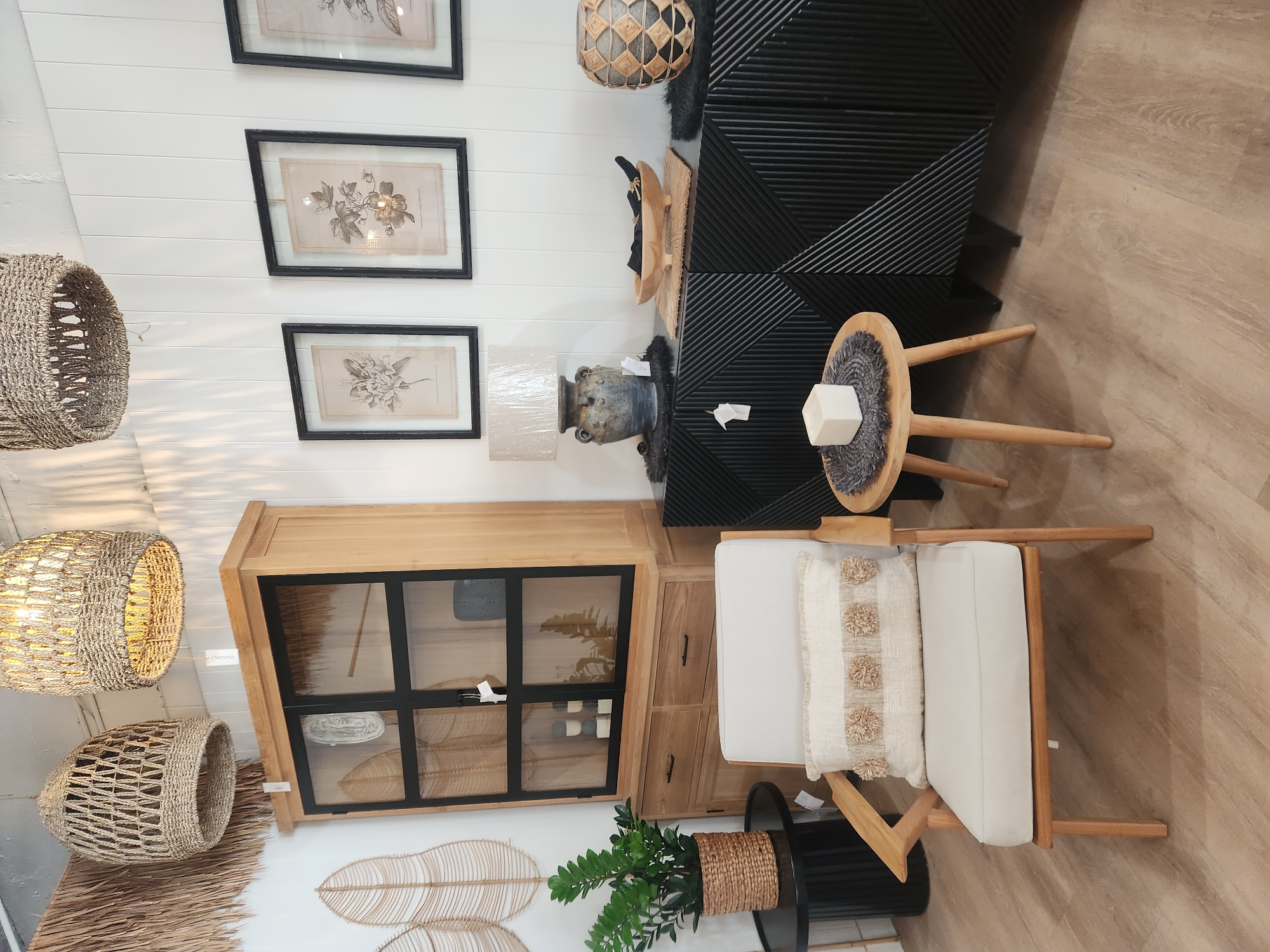 This image depicts a well-lit interior scene, likely a retail display or a staged room. The overall aesthetic is modern and natural, with a focus on wood tones, woven textures, and neutral colors.
In the foreground, a cream-colored armchair with a wooden frame is prominently featured. It is adorned with a decorative cushion featuring a cream background and textured beige pom-poms. To the left of the armchair, a black round side table holds a potted plant with lush green leaves.
To the right of the armchair, a small, light-wood tripod side table supports a white cube object placed on a dark grey woven mat. Behind this table is a large, black cabinet with a distinctive geometric pattern of diagonal lines etched into its surface.
Above the black cabinet, on a white paneled wall, are three framed black-and-white botanical prints. To the left of the prints hangs a wooden cabinet with glass doors, revealing shelves within. This cabinet has two drawers at the bottom.
Above the wooden cabinet, two large, woven pendant lights with a natural fiber texture are suspended from the ceiling. A third similar light is partially visible on the far left. The wall is white and paneled, providing a clean backdrop. The floor is a light-colored wood laminate.
There are no people visible in the image. The scene is static and appears to be set up for display or aesthetic purposes, showcasing furniture and decorative items. The location is not specified as Maitland, United States of America, and no such information can be inferred from the visual content.
