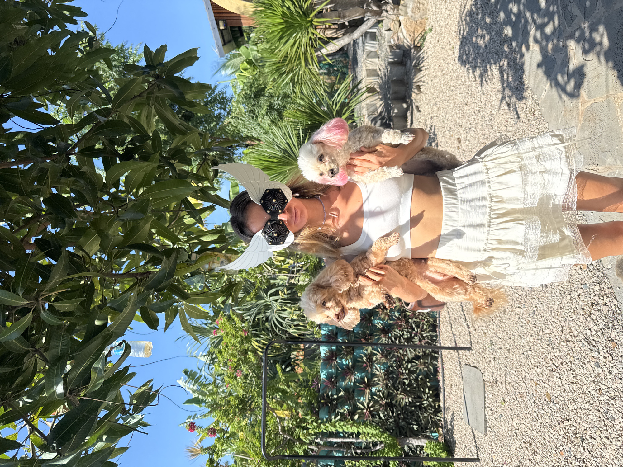 A woman in a white crop top and white skirt stands outdoors, holding two small dogs. She is wearing a white wing-shaped mask with black accents over her eyes. The first dog, a light brown poodle-mix, is held in her left arm. The second dog, a white poodle-mix with pink-dyed ears, is held in her right arm. The background features lush green foliage, palm trees, a blue sky, and a gravelly ground. The scene appears to be in a tropical or semi-tropical environment, consistent with locations like Mũi Né, Vietnam, known for its beaches and resort areas.