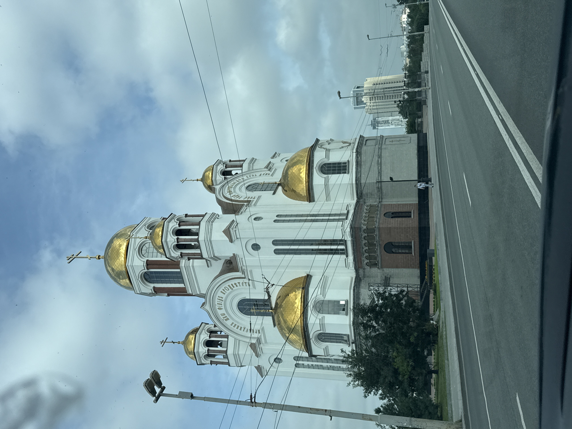 The image captures a frontal view of the Church on Blood in Yekaterinburg, Russia, under a cloudy sky. The church, a prominent landmark, features white walls with gold domes and crosses. In the foreground, a paved road with white lane markings is visible, suggesting it is a street. A few trees are present on the left side of the church. In the background, modern high-rise buildings are visible, indicating an urban setting. A solitary figure, appearing to be a person walking, is seen on the sidewalk to the left of the church. Utility poles and wires are also present, typical of a city environment. The overall scene is a daytime view of the church and its surroundings.