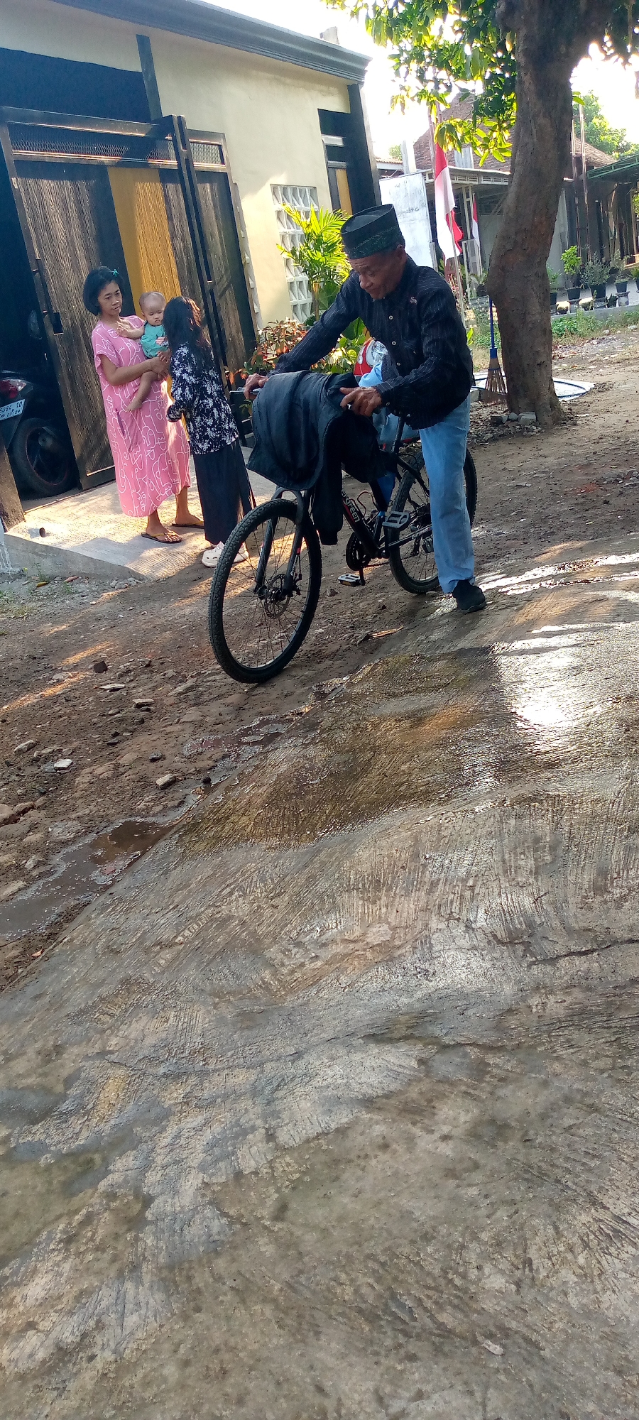 In Sumberwono, Indonesia, a man in a dark patterned shirt and a black cap is standing next to a bicycle with a black bag draped over the handlebars. He is wearing light blue jeans and dark shoes. To his left, a woman in a pink dress is holding a baby, and a young girl in a black and white patterned outfit stands beside her. The group is near the entrance of a building with a large wooden door. The ground in front of them is a mix of dirt, gravel, and a wet concrete surface. A tree with green leaves is visible to the right of the man, and a red and white flag is mounted on a pole in the background. Sunlight casts shadows across the scene.