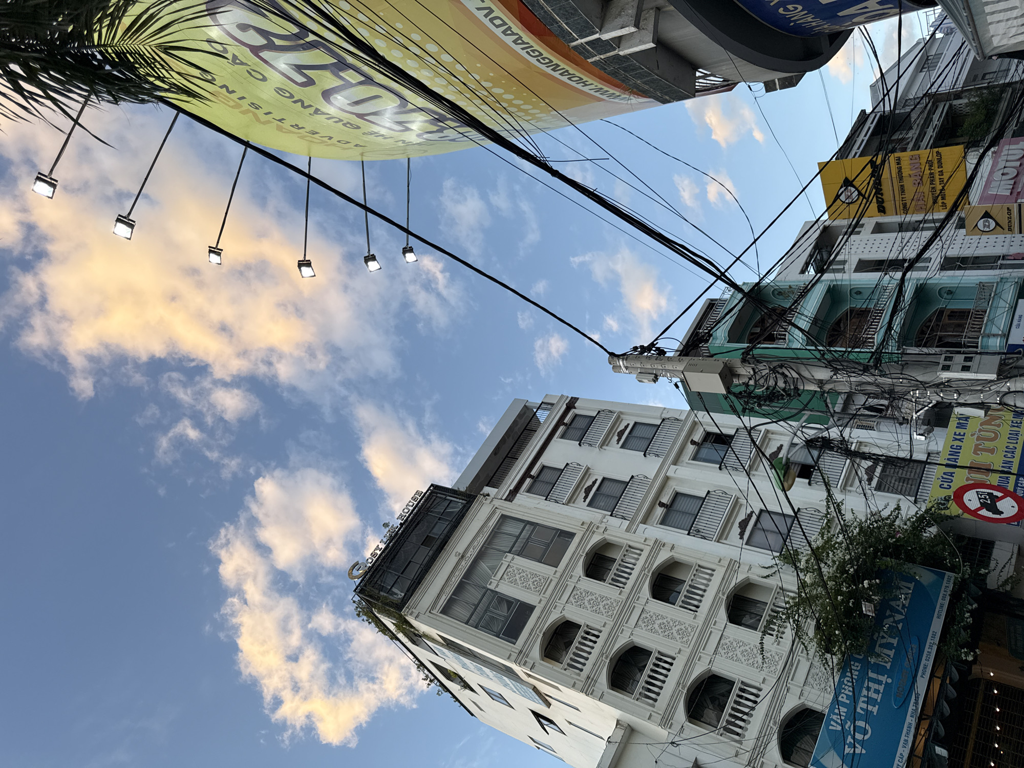 The image, taken in Nha Trang, Vietnam, captures a street scene dominated by a modern white building and a large curved billboard. The white building, with multiple stories featuring balconies and ornate architectural details, is positioned to the left of the frame, leaning slightly. Atop this building, a sign reads "LOST IN HOUSE". To the right, a large, yellow, curved billboard advertising "HOANG GIA ADV" is prominently displayed, illuminated by a series of evenly spaced spotlights. The sky above is a bright blue with scattered, wispy clouds, some tinged with yellow light. A dense network of black electrical wires crisscrosses the foreground, creating a visually complex overlay on the buildings and sky. Several smaller signs and storefronts are visible at the base of the buildings, indicating commercial activity. One sign in Vietnamese reads "CUA HANG XE MAI" (motorcycle shop) with the brand "MOTUL" also visible. Another blue sign at the bottom left reads "VỞ THỊ MỸ NAM" which translates to "Vo Thi My Nam Office." The perspective is upward-looking, emphasizing the height of the buildings and the expanse of the sky.