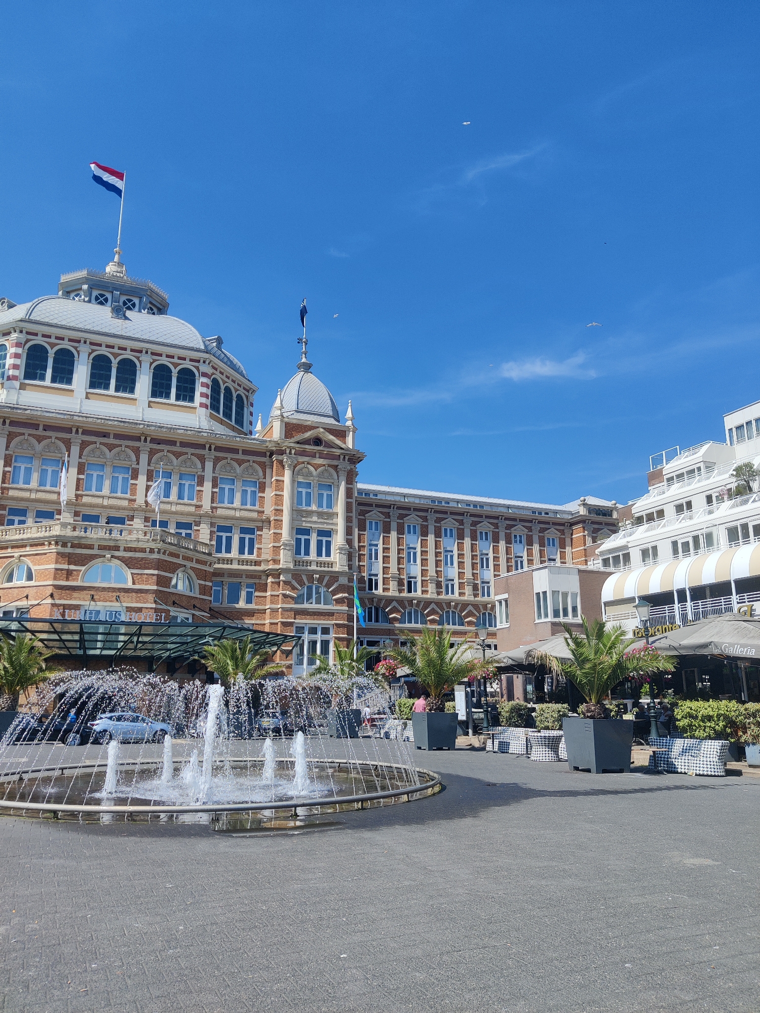 The image captures the exterior of the Kurhaus Hotel in Scheveningen, The Hague, Netherlands, on a clear, sunny day. In the foreground, a circular fountain with multiple jets of water is active, spraying water into the air and creating a mist. The ground around the fountain is paved with grey interlocking bricks.
Behind the fountain, several palm trees are planted in large, dark grey square planters. Outdoor seating areas for cafes and restaurants are visible, featuring tables and chairs. Some of the seating is covered by umbrellas, and checkered cushions are placed on some of the chairs. The signage on one of the awnings reads "La Galleria".
The Kurhaus Hotel itself is a large, ornate building with a distinctive red-brick facade and white architectural details. It features multiple stories with numerous windows, a domed roof section, and another smaller dome topped with a flag. The Dutch flag is prominently displayed on the highest dome. The hotel's entrance area has a glass canopy. To the right of the Kurhaus, a modern, white multi-story building with balconies and glass facades is visible.
The sky is a vibrant blue with a few wispy clouds and several birds flying in the distance. A few people are present, some sitting at the outdoor tables, and others are likely passing by or working near the hotel entrance. The overall scene depicts a bustling public space adjacent to a historic hotel, likely a popular tourist destination in the Scheveningen seaside resort.