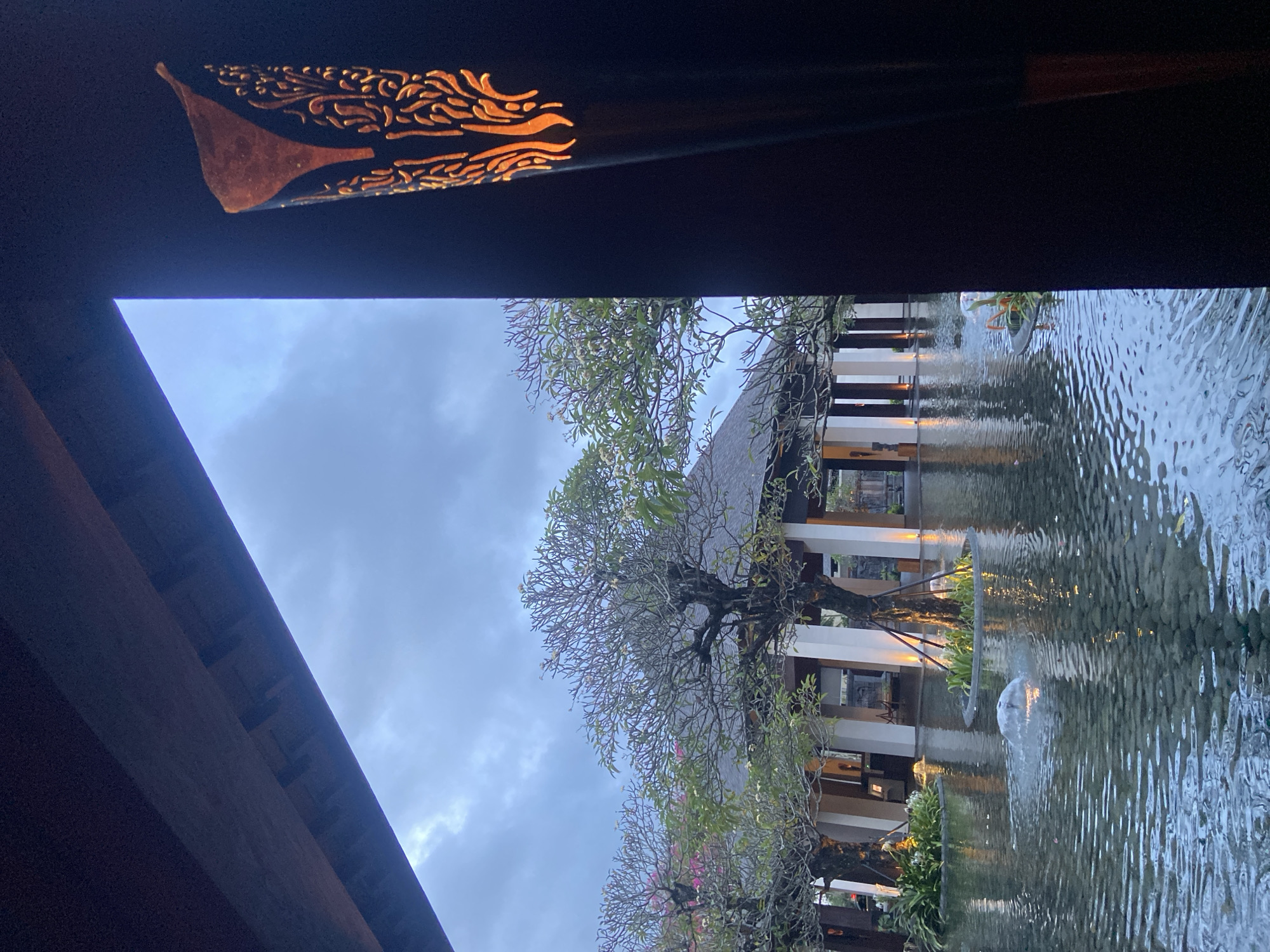 The image shows a view framed by a dark, ornate structure, likely part of a building. Beyond the frame, there's a courtyard with a building featuring white columns, reflected in a body of water, likely a pond or pool. Trees with bare branches and some sparse foliage are situated between the building and the water. In the water, a fountain is visible. The sky is overcast with grey clouds. The location is Kuta Selatan, Indonesia.