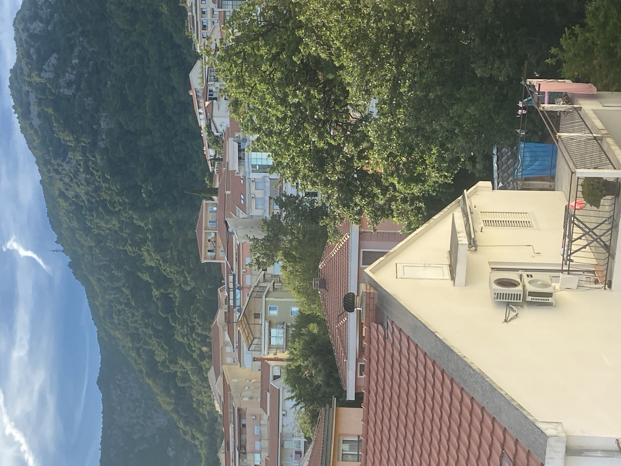 The image depicts a view of the city of Budva, Montenegro, as seen from an elevated position. In the foreground, the roof and upper portion of a light-colored building are visible, featuring a red-tiled roof and an attached air conditioning unit. A balcony with a railing is also present, with laundry hanging on a clothesline.
Behind this building, a dense cluster of residential buildings with red-tiled roofs fills the mid-ground, exhibiting various architectural styles and colors, including cream, yellow, and light pink. Trees interspersed among the buildings add greenery to the scene.
Dominating the background is a lush, green mountainside covered in dense vegetation. The sky above is a mix of blue and white, with wispy clouds and what appears to be a distant electrical tower. The overall impression is a residential area nestled against a natural, mountainous backdrop, characteristic of coastal towns in the region.