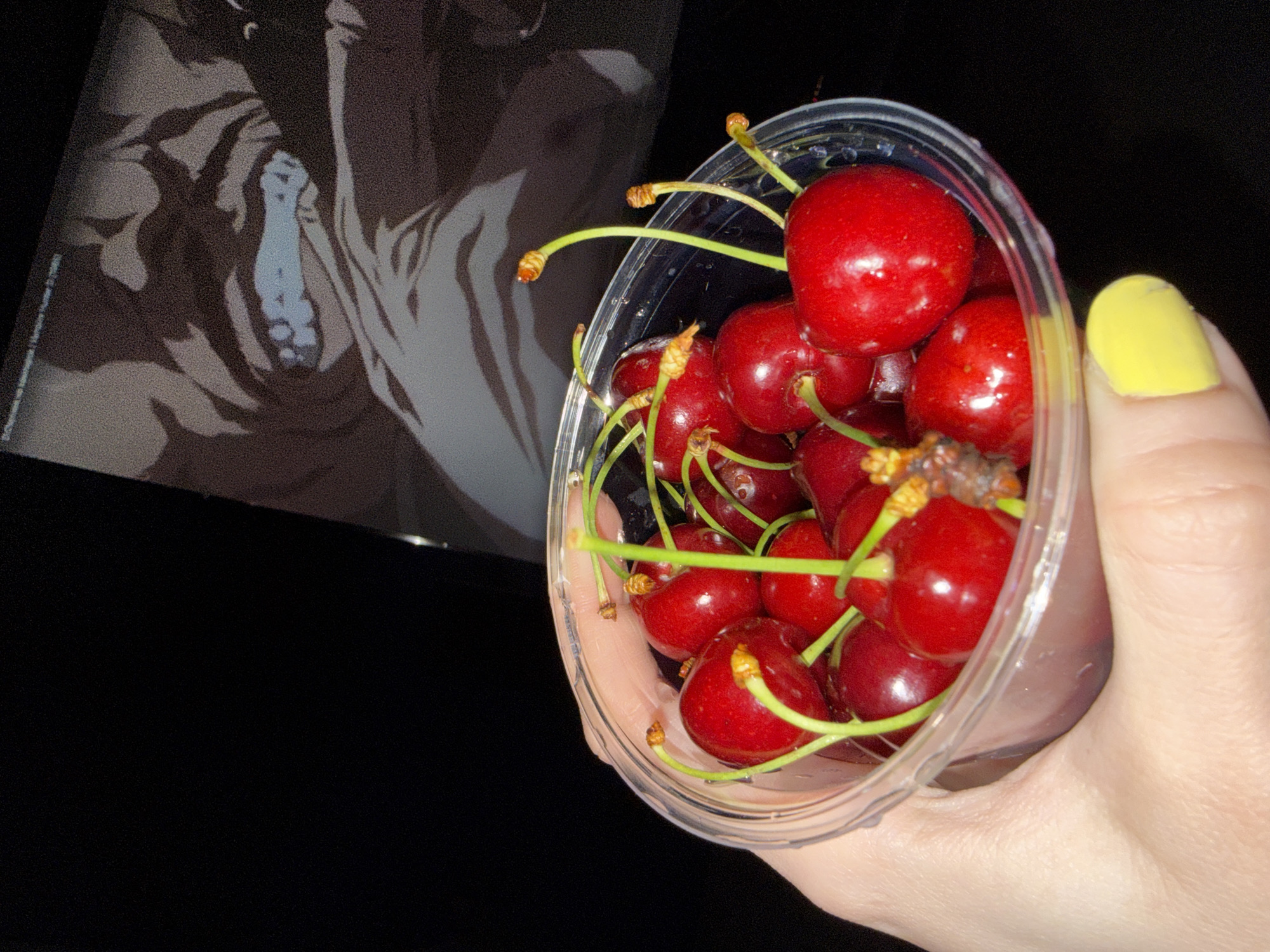 A hand, with a yellow painted fingernail, holds a clear plastic container filled with fresh red cherries with green stems. The background is dark, with a blurry image of a stylized figure in shades of brown and white visible to the left of the container. The location in the city of Moscow, Russia is unknown.