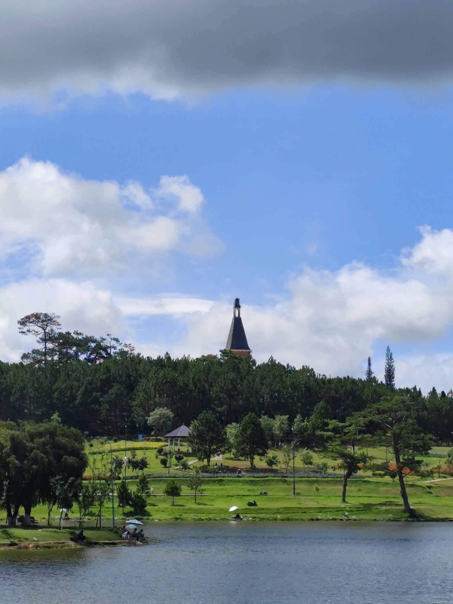 The image depicts a tranquil scene in Da Lat, Vietnam, likely near Xuan Huong Lake. The lake occupies the lower portion of the frame, its surface reflecting the sky. In the foreground, figures are seated along the shoreline under umbrellas, possibly fishing or relaxing. A grassy bank leads up to a park with walking paths, various trees, and a gazebo in the midground. A dense line of trees obscures the horizon. A tower structure, reminiscent of European architecture, is visible above the trees. The sky features a combination of blue sky and scattered clouds.