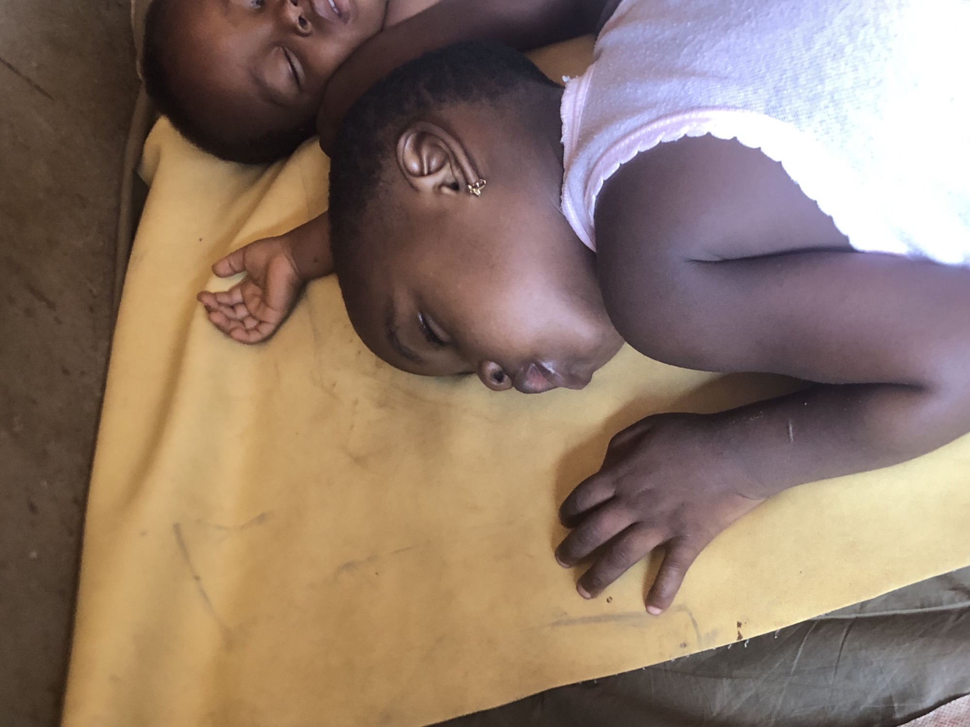 Two young Black children are sleeping on a yellow cloth, possibly a mat or bedsheet, in what appears to be an outdoor or semi-outdoor setting in Ukwu-Abwa, Nigeria. The child in the foreground is lying on their side, facing away from the camera, with their head turned towards the viewer. They are wearing a light pink top with a scalloped edge. A small, gold-colored earring with a butterfly design is visible in their left ear. Their right arm is bent at the elbow, with their hand resting on the yellow cloth, fingers slightly curled. The other child is partially visible behind the first, also sleeping. Their head is turned towards the camera, and their eyes are closed. The lighting suggests it is daytime, possibly late morning or early afternoon, with bright natural light illuminating the scene. The background is somewhat blurred, but appears to be a rough, earthy surface, consistent with an outdoor environment in a rural or developing area. The overall mood is peaceful and serene, capturing a moment of rest for the children. There is no visible text in the image.
